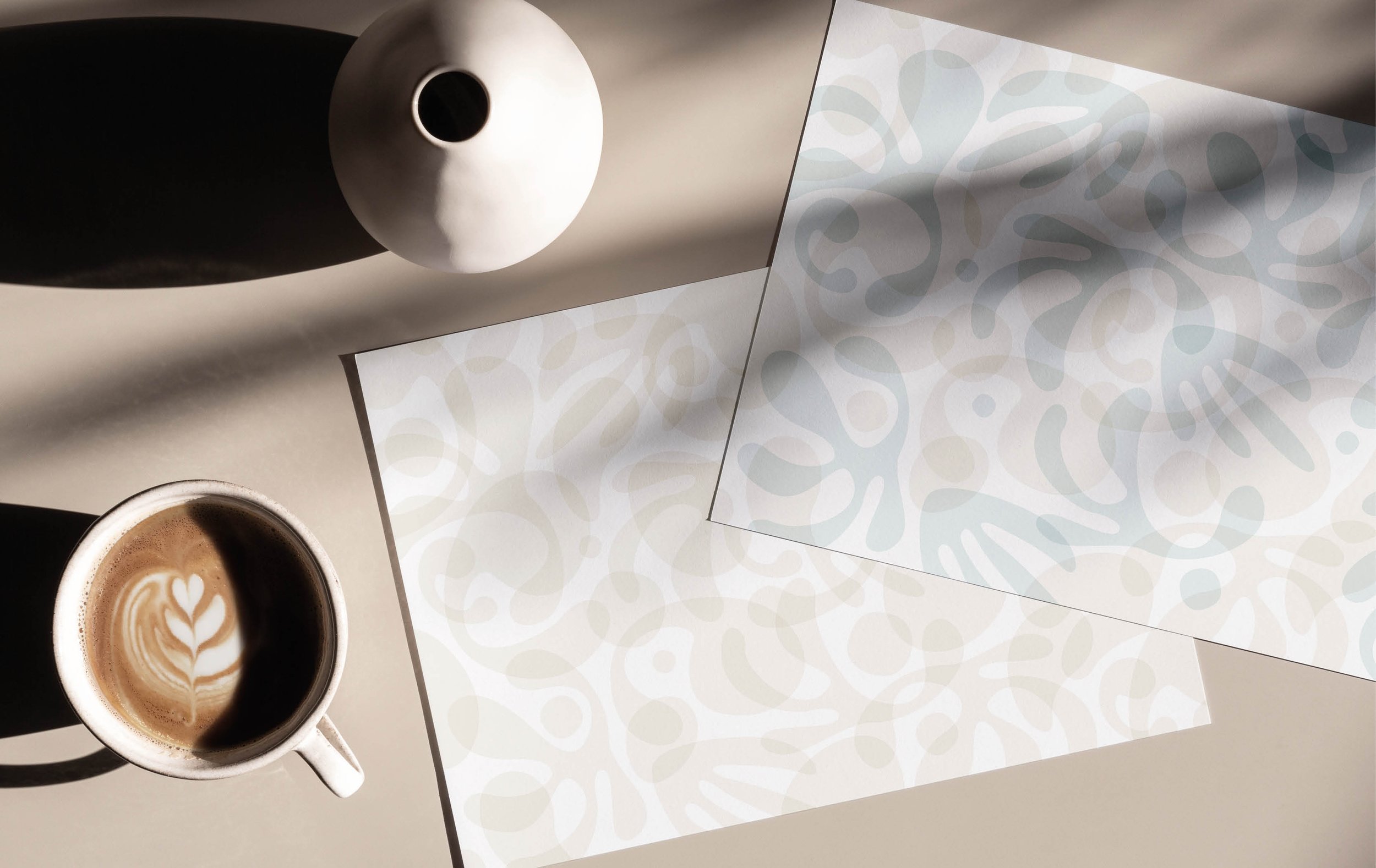 A cup of coffee with latte art, a white bowl with a black interior, and a decorative sheet of paper with a light blue abstract pattern on a flat surface.