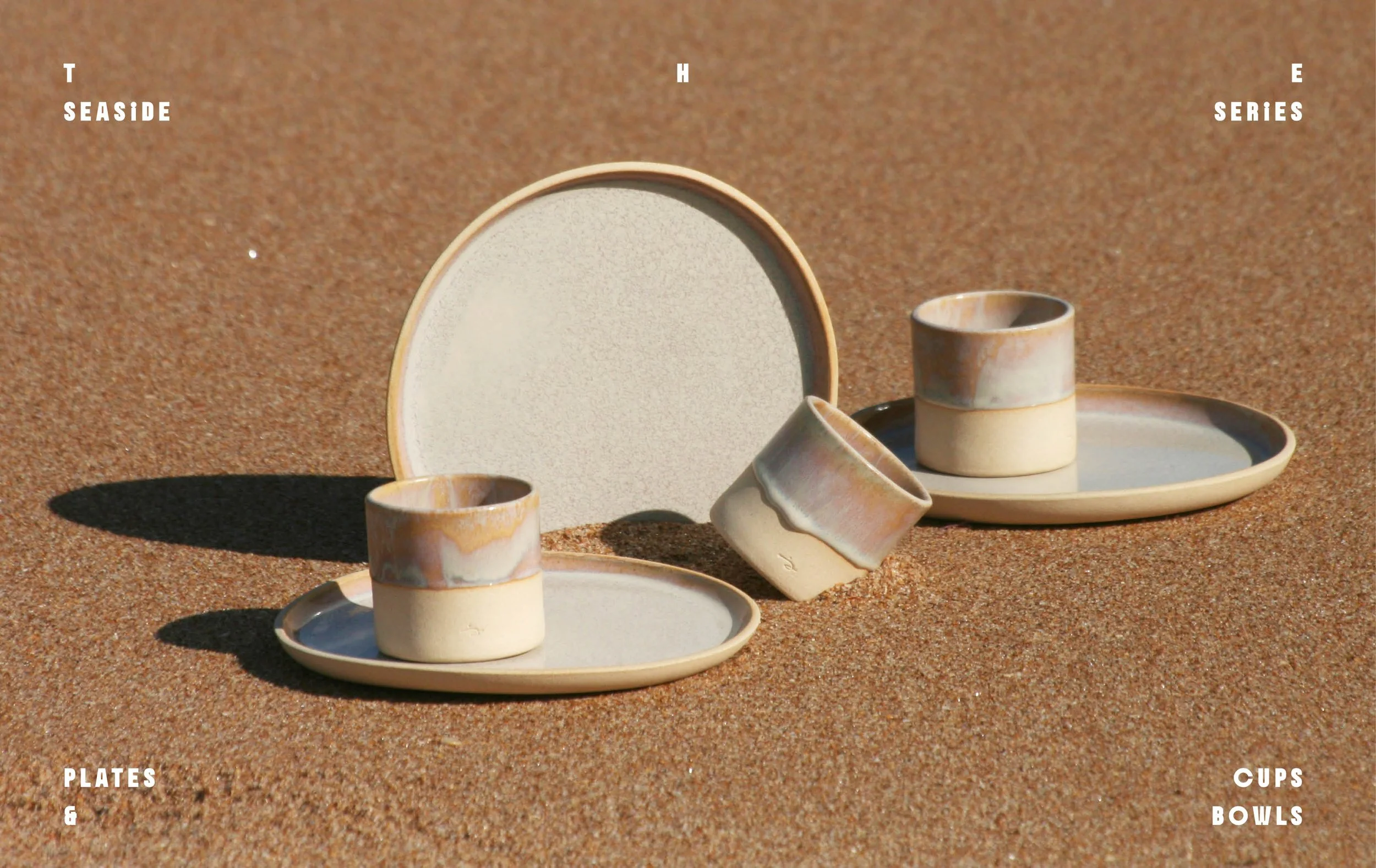 A set of beige and pastel-colored ceramic plates, bowls, and cups arranged on a sandy surface with shadows cast by sunlight