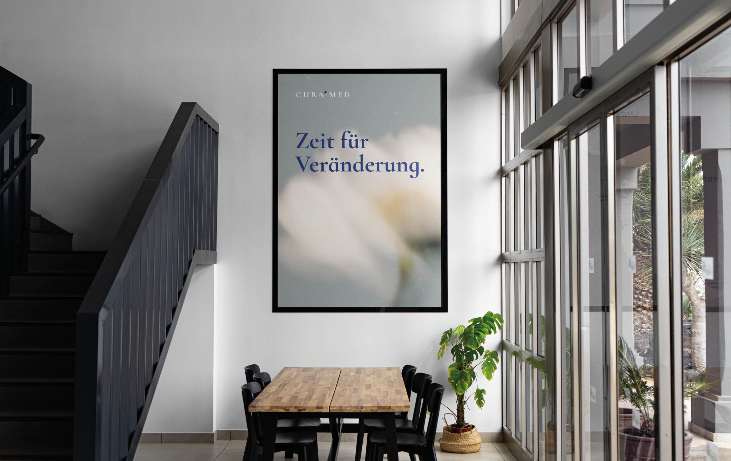 Modern interior space with a staircase on the left, a framed wall art with German text 'Zeit für Veränderung' at the center, large glass windows on the right, a wooden dining table with black chairs, and a potted plant near the window.
