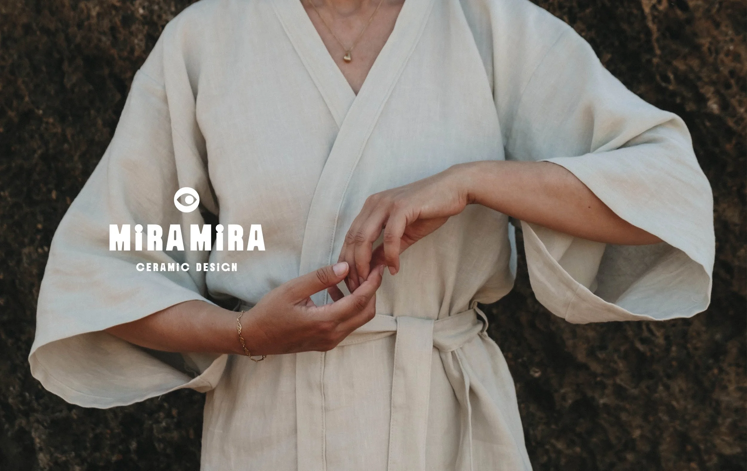 Close-up of a person wearing a beige traditional kimono or robe, holding their hands together in front of their waist, with a background of dark soil or rocks.