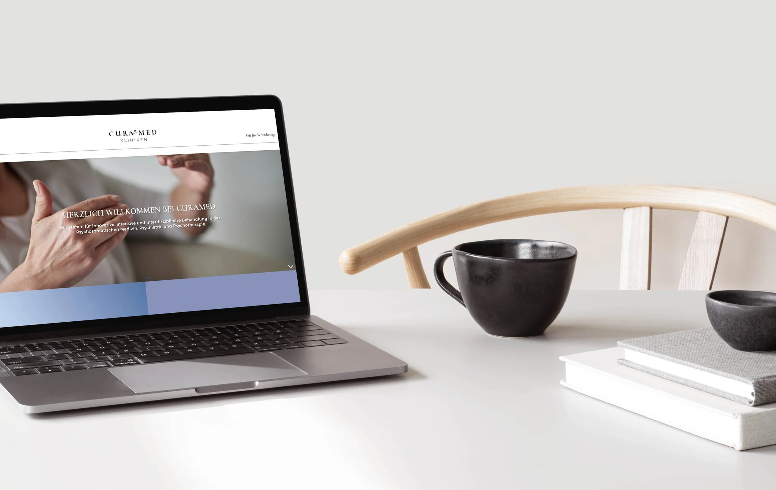 Part of a laptop displaying a website in German, on a white desk with black and gray ceramic cups, and a few stacked books with gray and white covers, against a light gray wall.