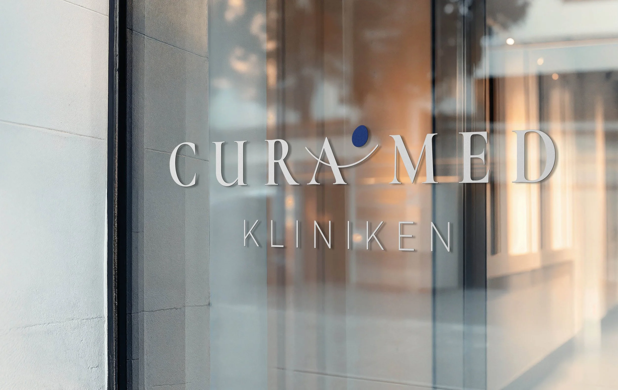 Glass door with the words 'CURED MED' and 'KLINIKEN' written on it, reflecting the interior of a modern clinic or medical facility.