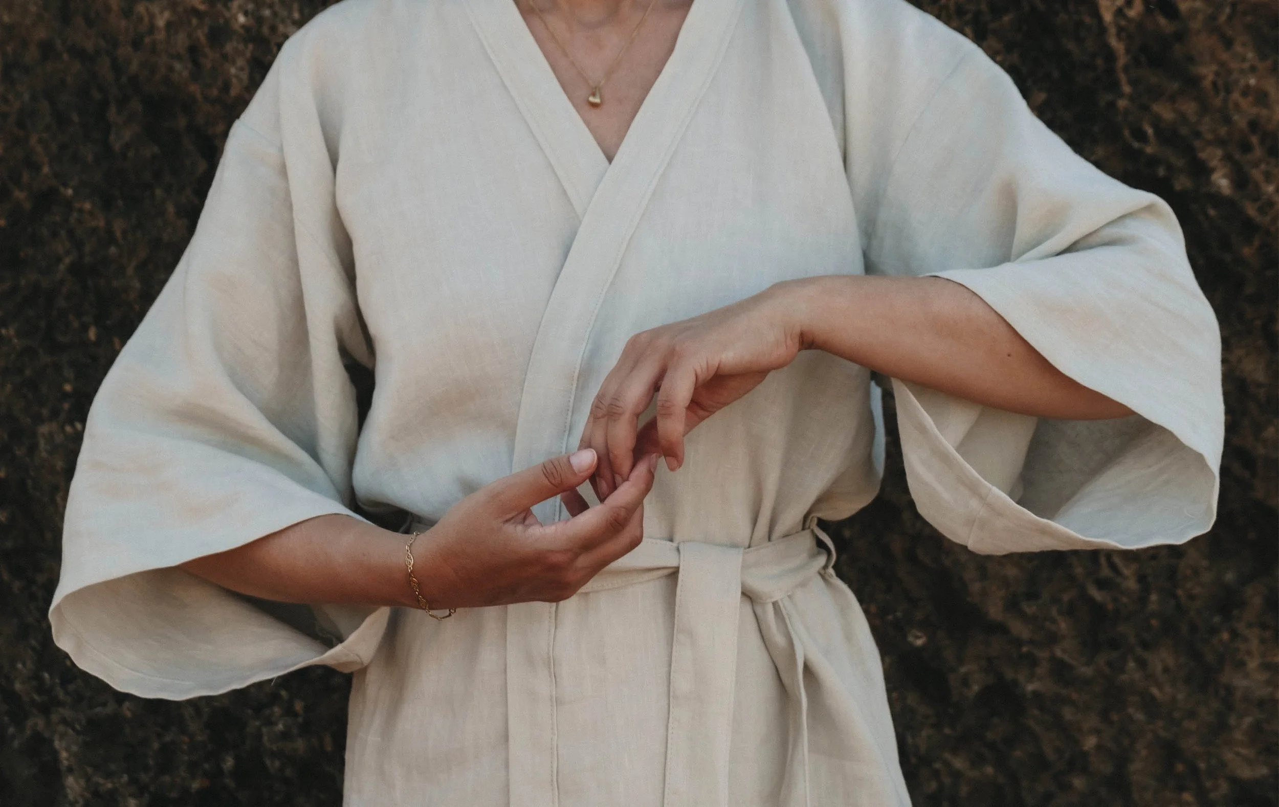 A person wearing a white kimono-style robe with a belt, standing on a dark textured surface, adjusting their wrist with a gold bracelet and wearing a gold pendant necklace.