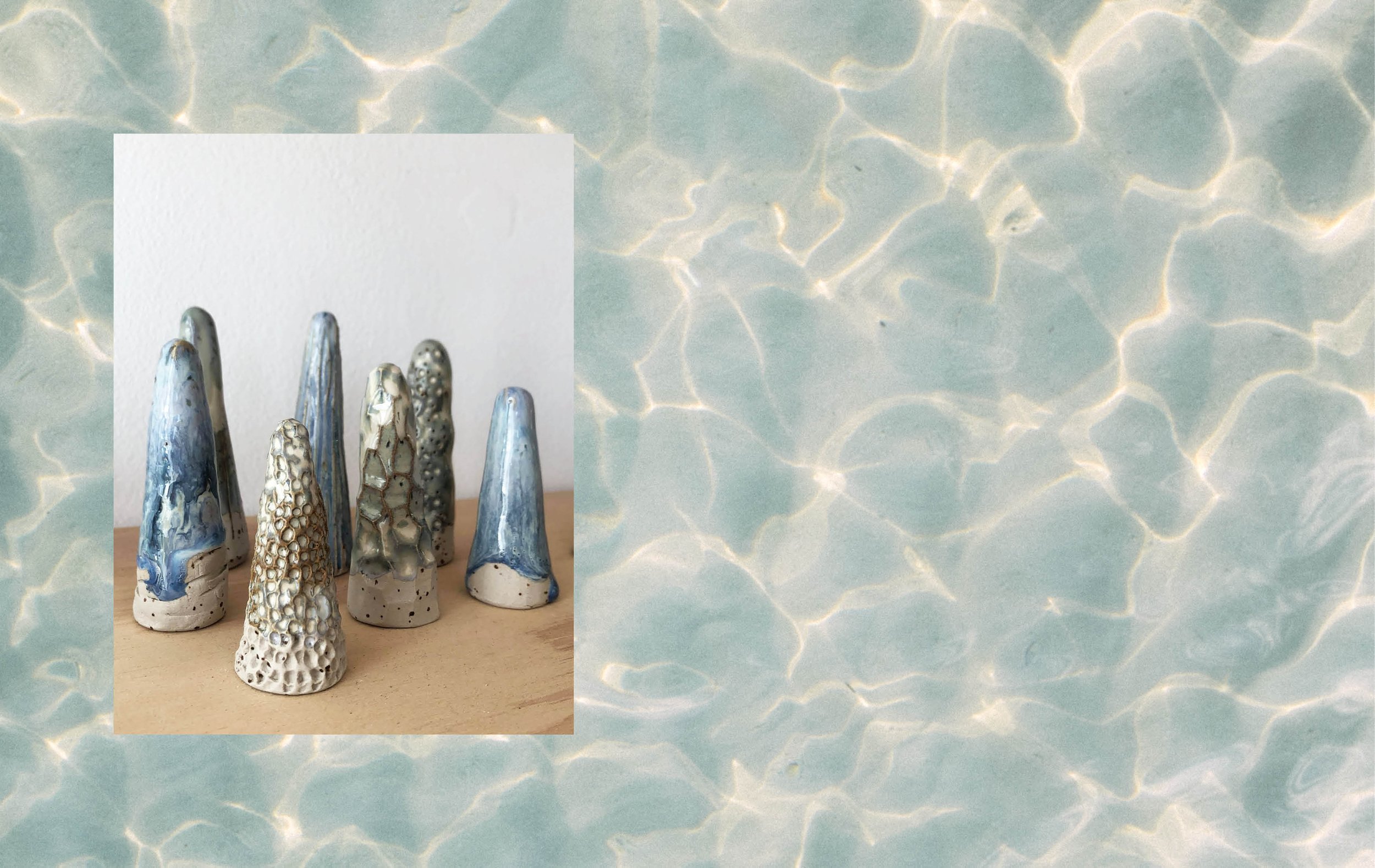 A set of ceramic sculptures resembling fingers, each with unique textures and shades of blue, cream, and gray, displayed on a wooden surface against a background of water-reflection patterns.