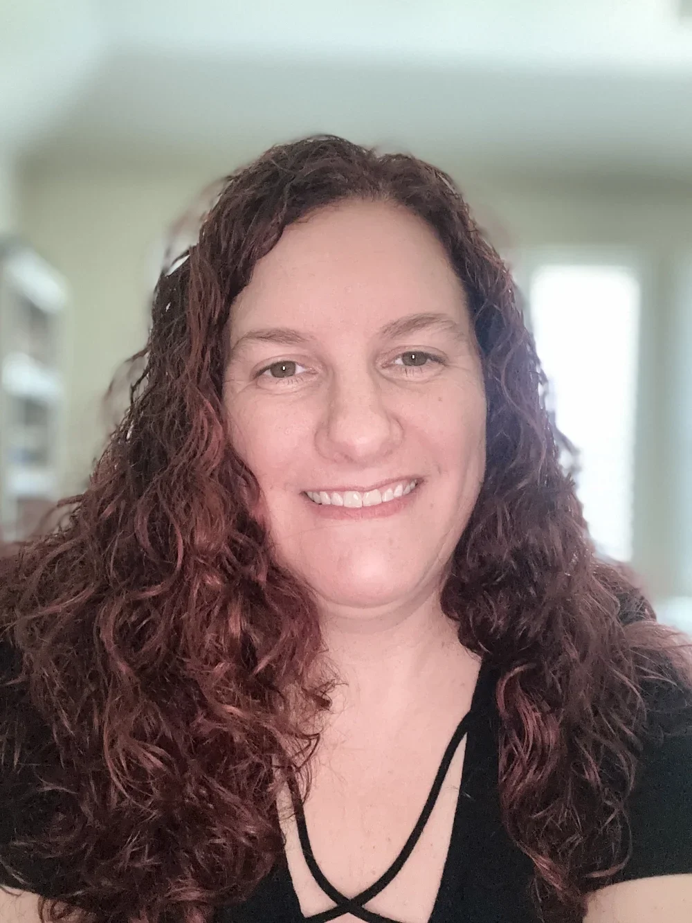 A woman with curly red hair smiling, inside a home with a blurred background of a room with light coming through windows.