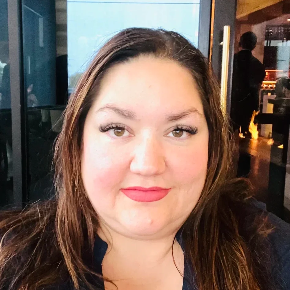 Close-up of a woman with long brown hair and light makeup, smiling at the camera, inside a restaurant or cafe with dim lighting and a person in the background.