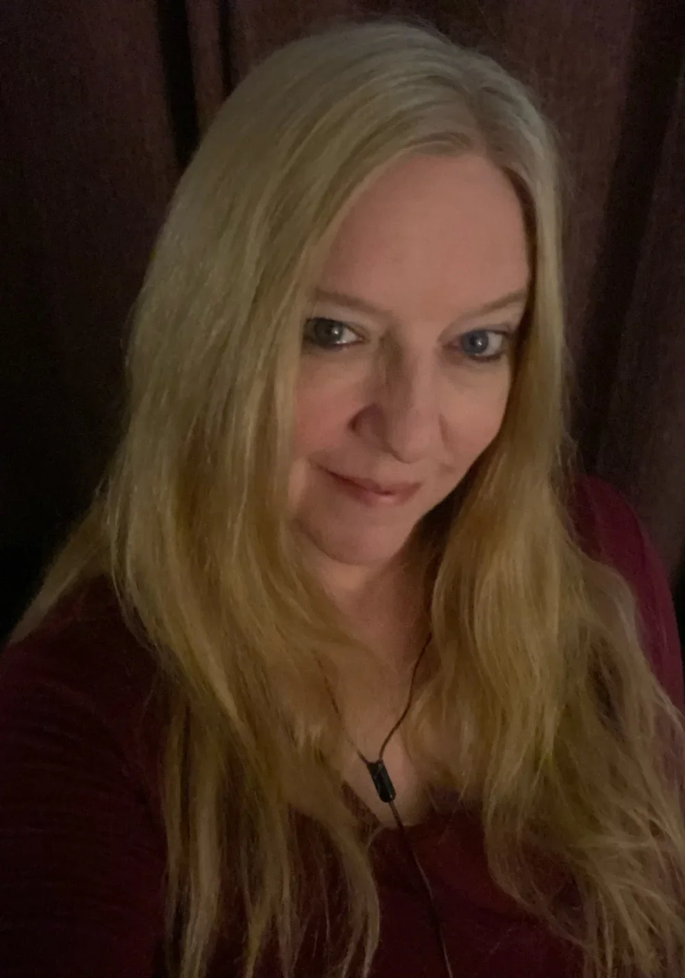 A woman with long blonde hair and blue eyes smiling slightly, wearing a dark red top and a black necklace, standing against a dark brown wooden background.