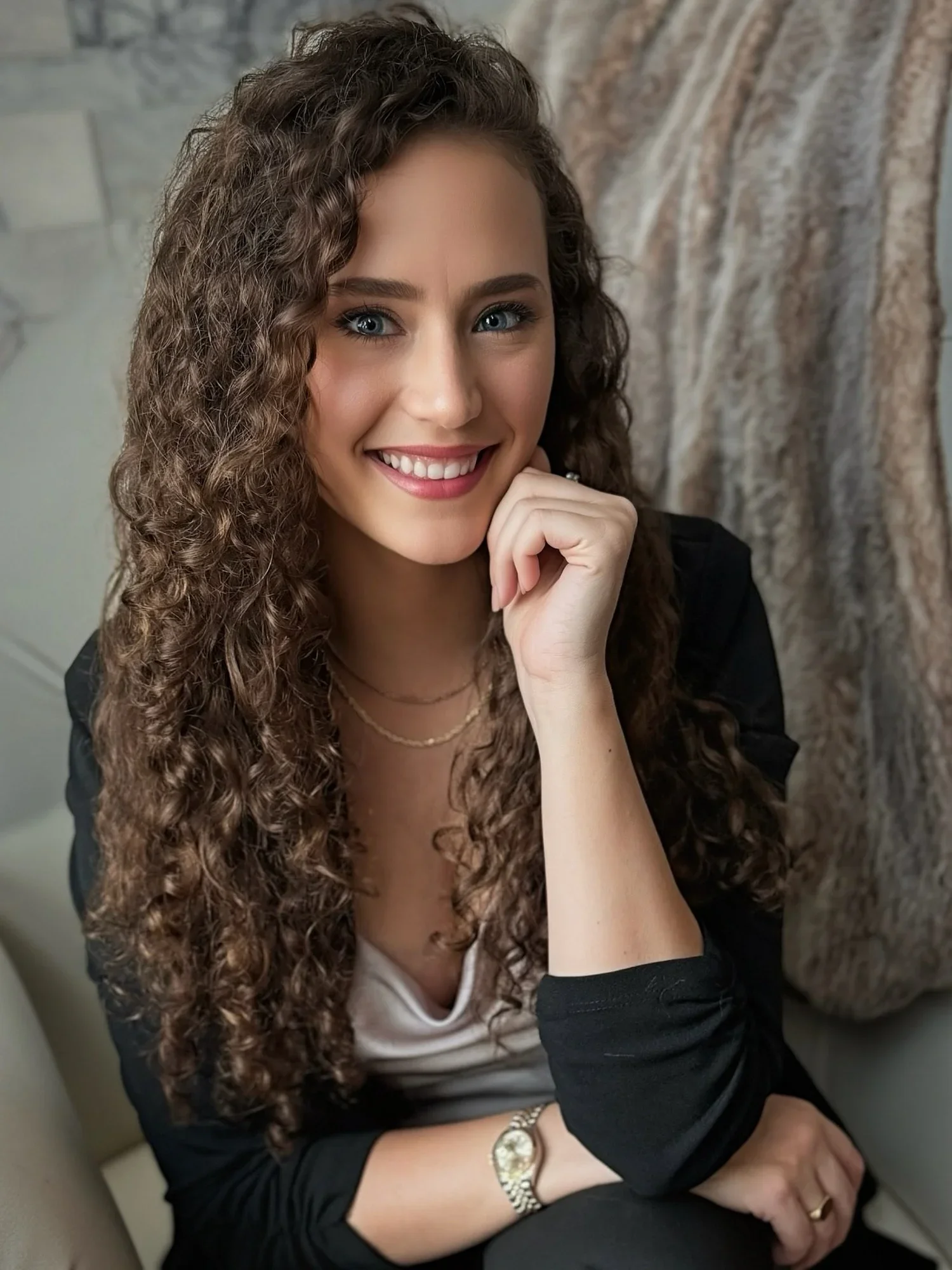 A young woman with curly brown hair and blue eyes smiling, resting her chin on her hand, sitting on a light-colored chair with a curtain in the background.