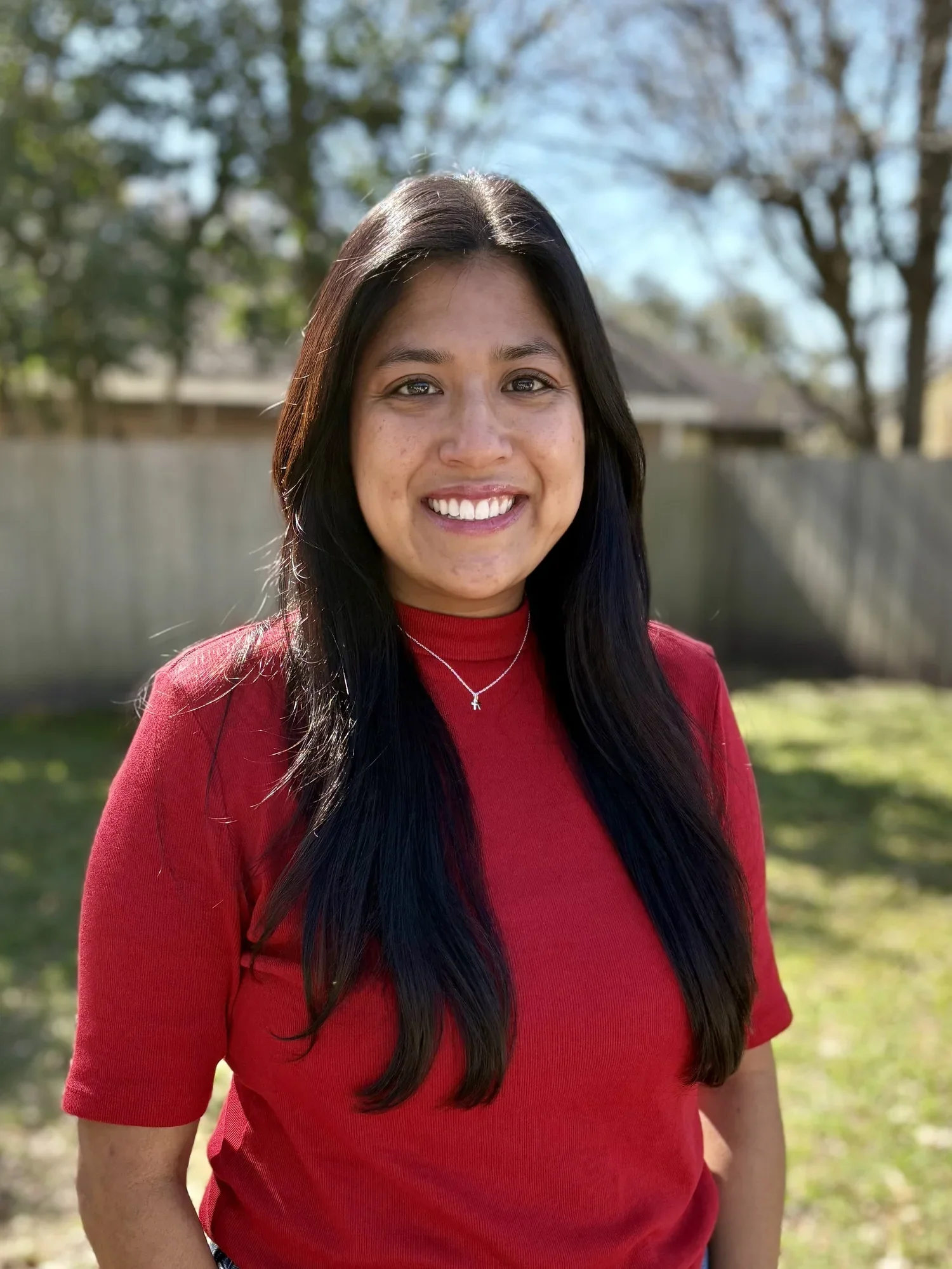 A young woman with long dark hair wearing a red top and a delicate necklace, smiling outdoors with a fence and trees in the background.