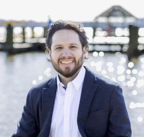 A man with dark hair and a beard smiling outdoors near water, wearing a white shirt and navy blazer, with a pier and boat house in the background.
