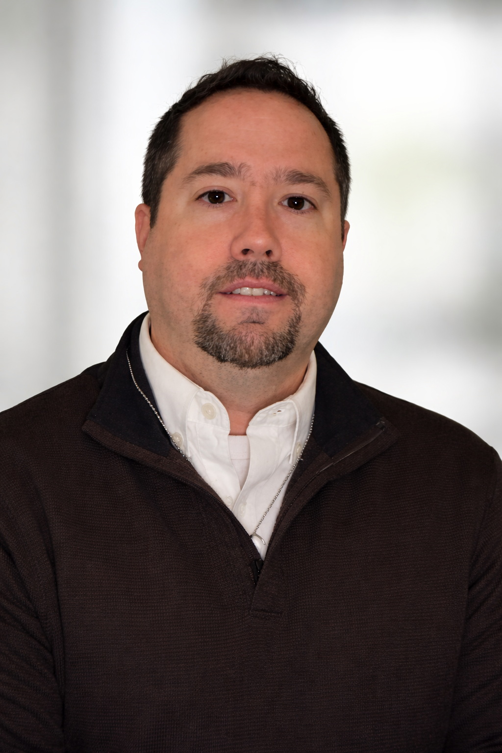 A close-up portrait of a man with short dark hair and a goatee, wearing a white collared shirt and a black zip-up jacket, standing against a blurred light-colored background.