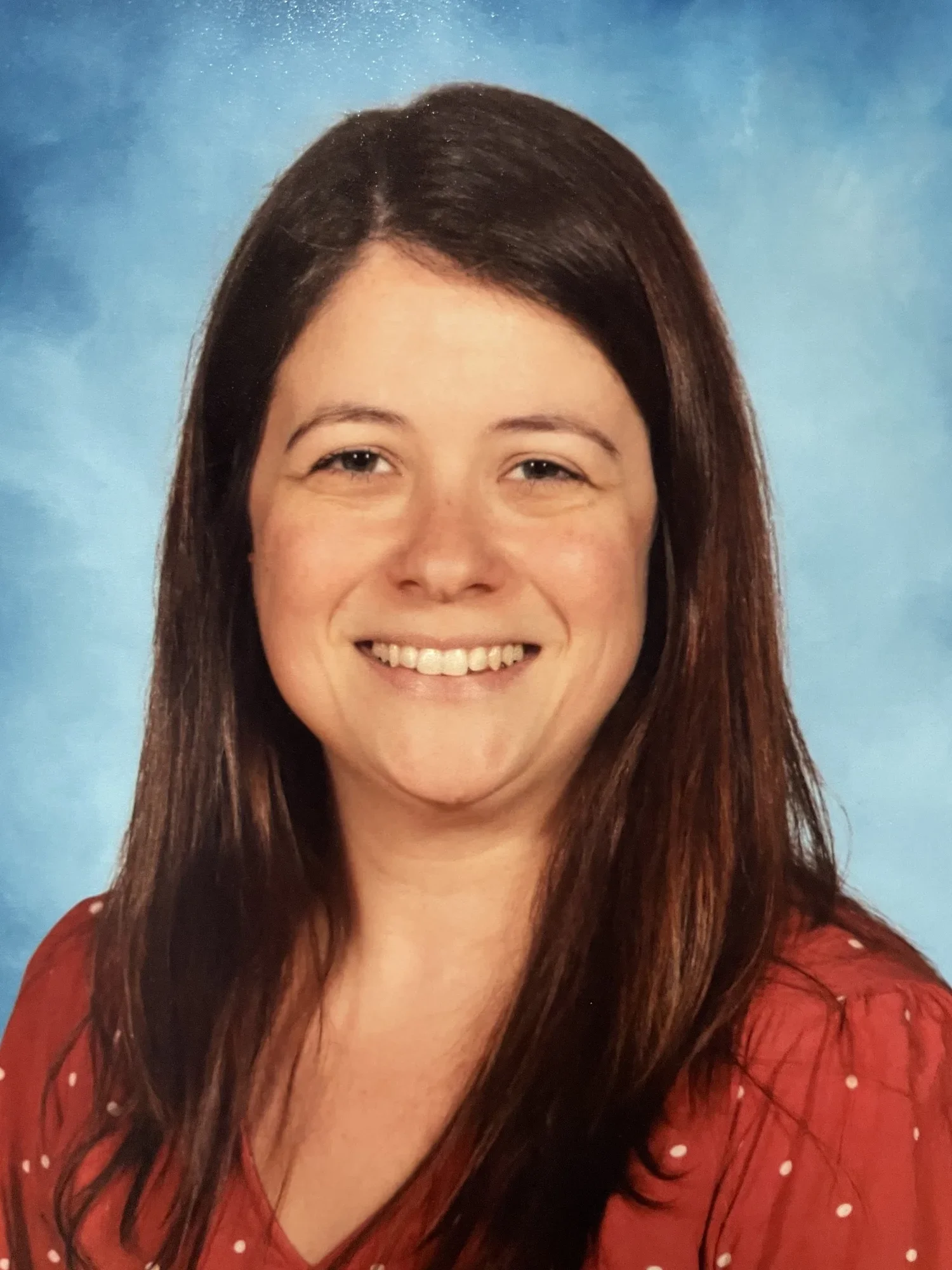A woman with shoulder-length dark brown hair smiling against a blue background, wearing a red top with white polka dots.