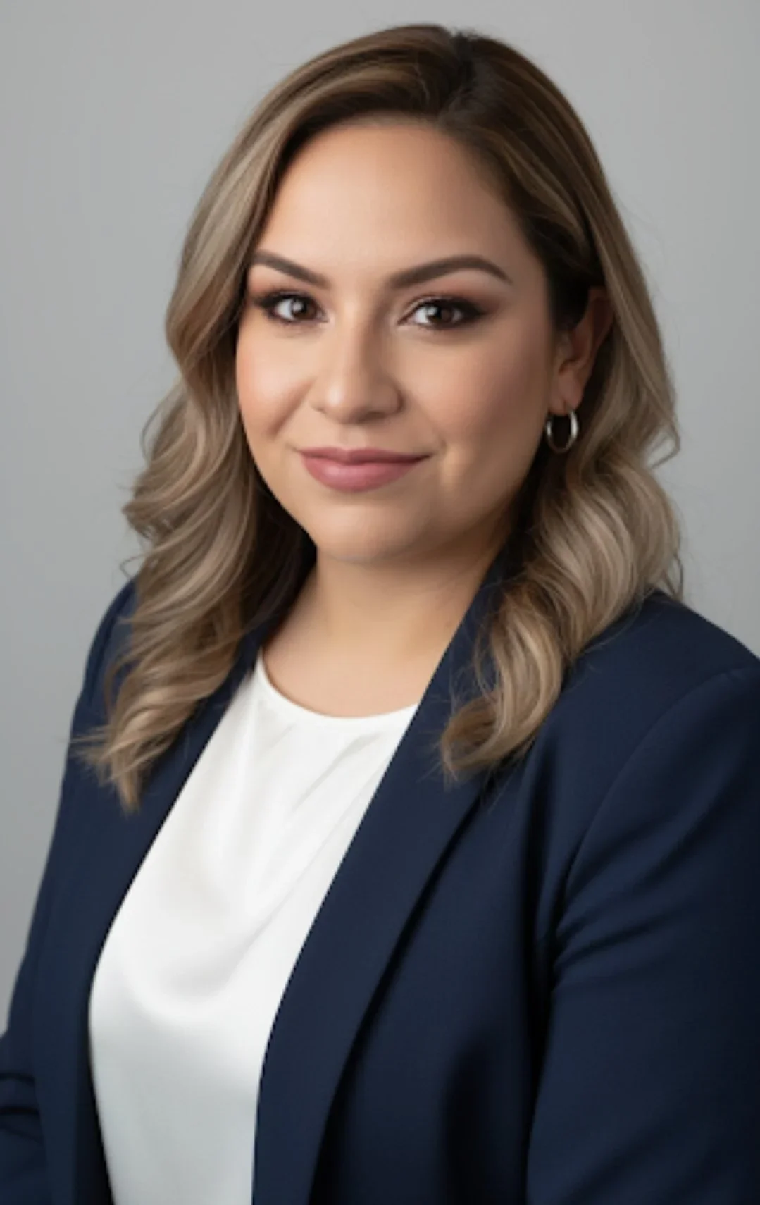 A professional woman with wavy, shoulder-length light brown hair, wearing a navy blazer and a white top, standing against a plain gray background.