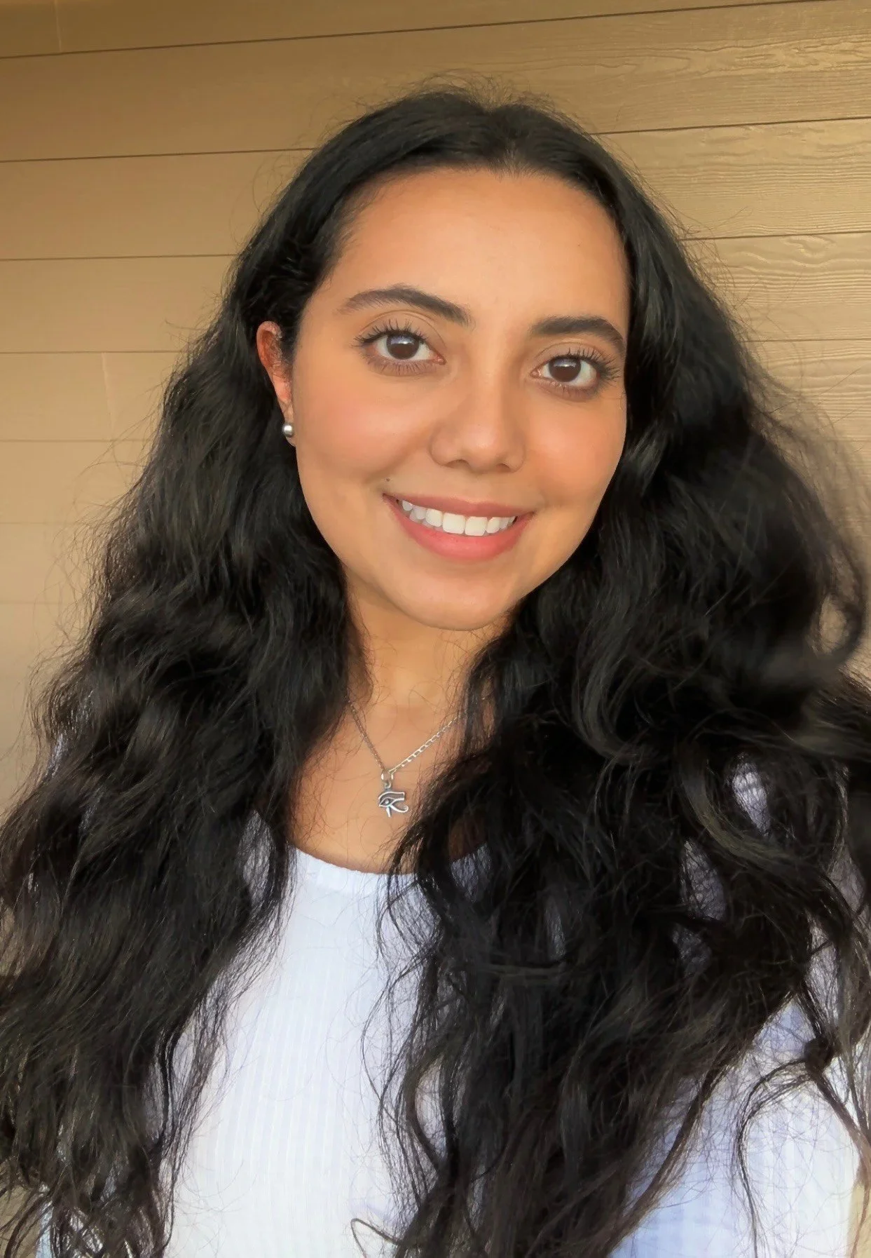 Close-up of a woman with long, wavy black hair, wearing pearl earrings, a necklace with an eye symbol, and a white top, smiling in front of a wooden wall background.