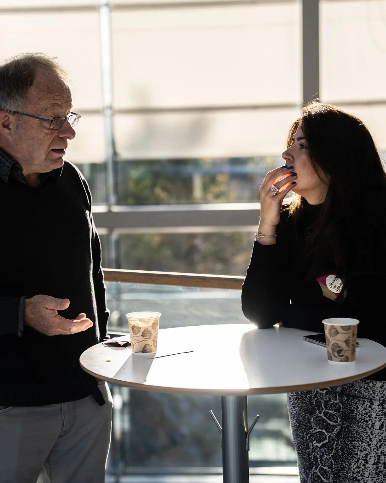 A man and woman engaged in conversation at a round table with paper cups, in a bright indoor setting with large windows.