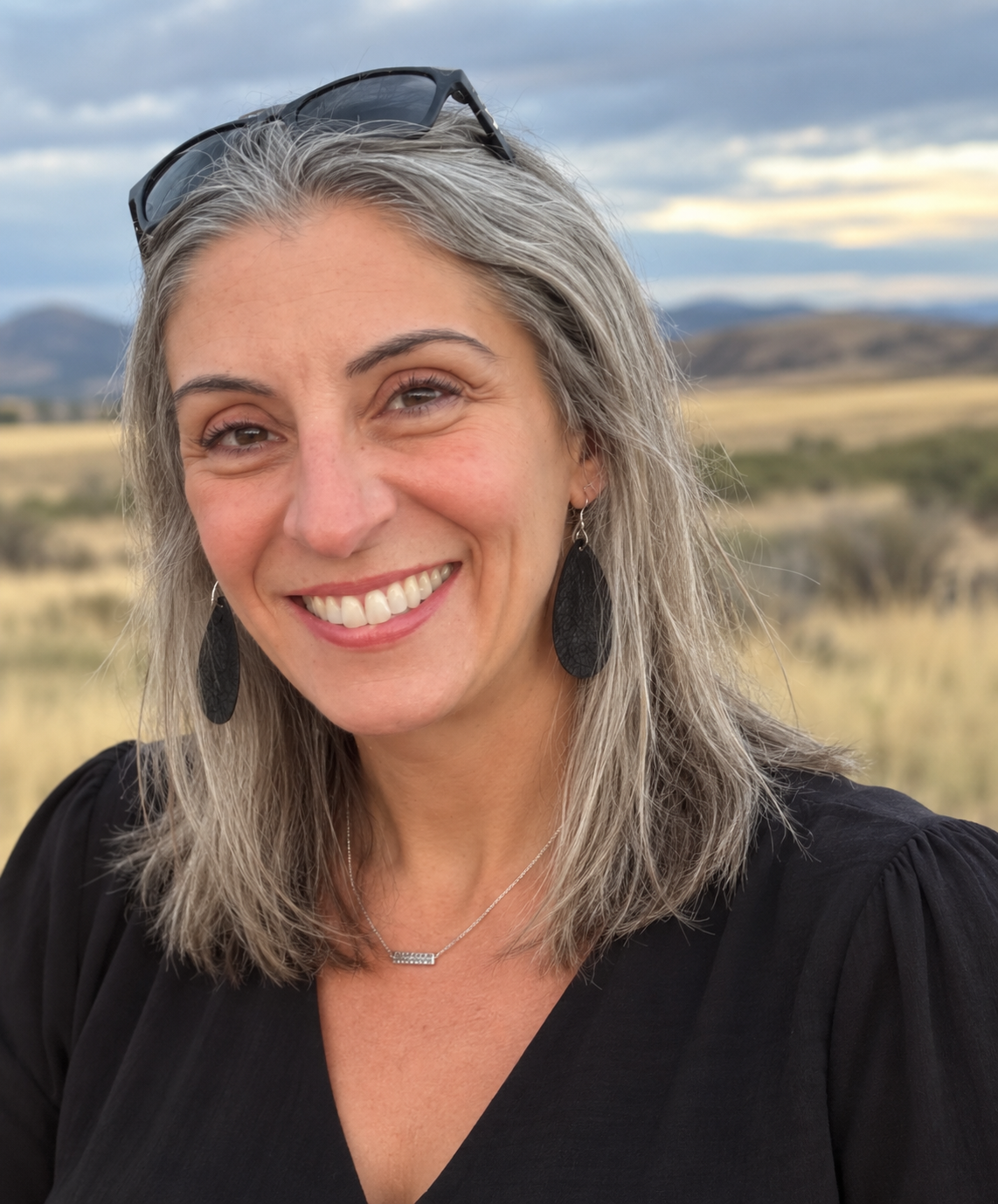 A woman with gray hair, wearing glasses on her head and black earrings, smiling outdoors with a grassy landscape and mountains in the background.