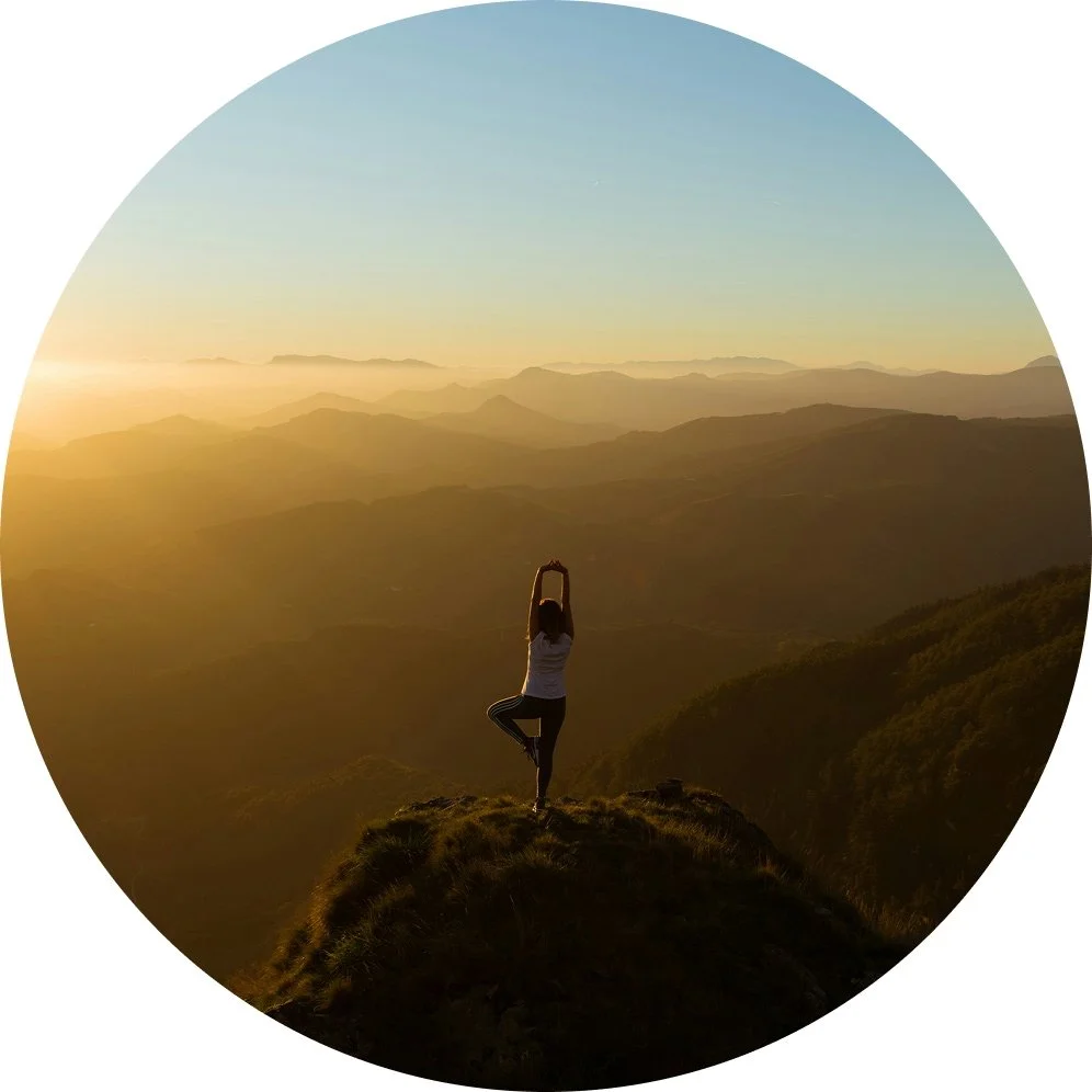 Person standing on a mountain peak in a yoga pose during sunrise, overlooking a landscape of rolling hills and mountains.