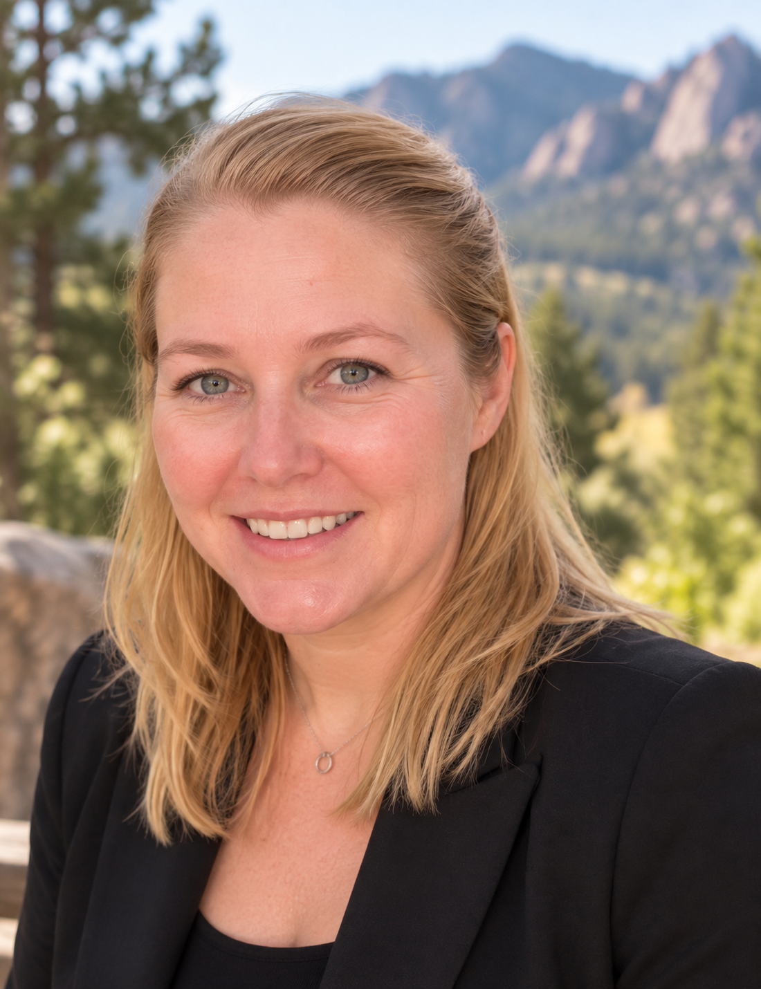 A woman with blonde hair smiling outdoors with mountains and trees in the background.