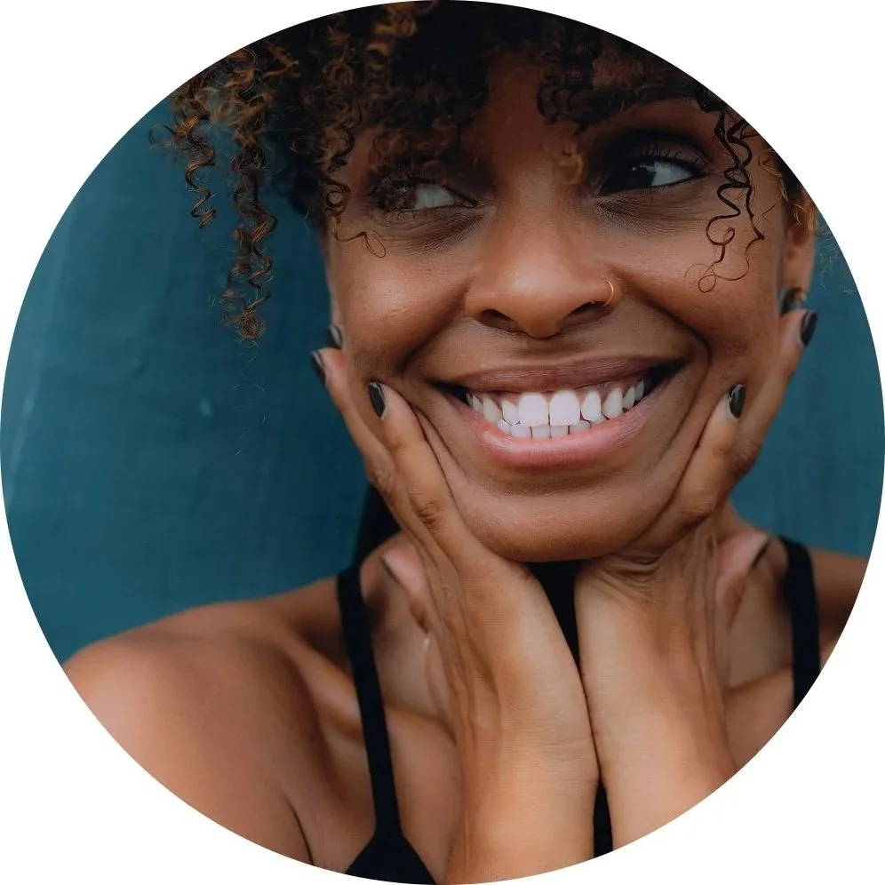 Close-up of smiling woman with short curly hair, nose ring, and black nail polish, holding her face with both hands and looking slightly to her right.