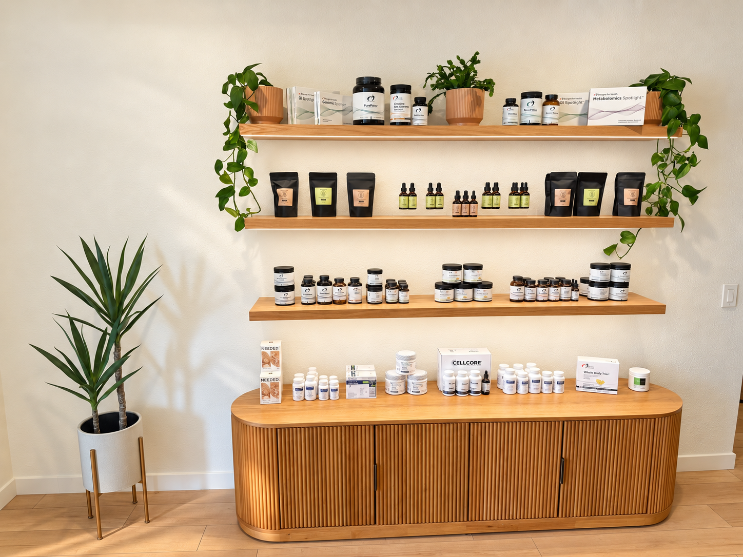 Display of health supplements and products on wooden shelves and a cabinet, with potted plants on top and to the side, against a beige wall.