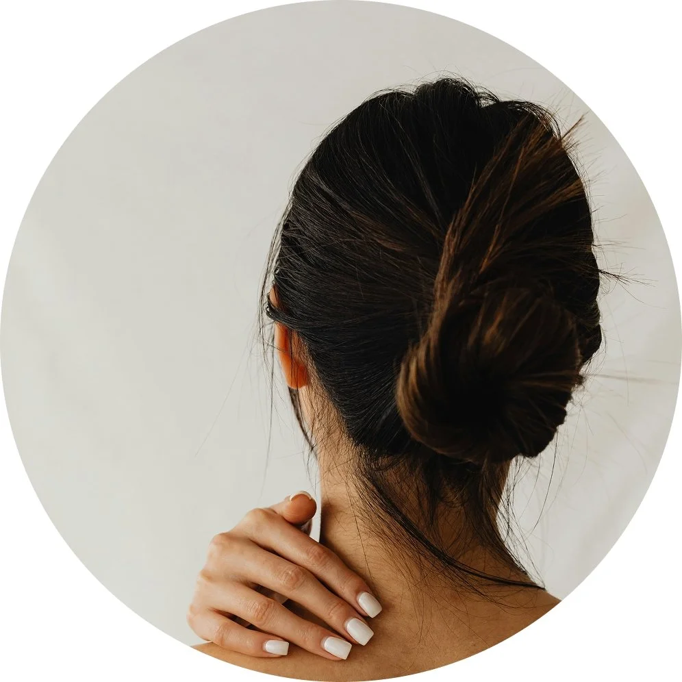 Back view of a woman with dark hair styled in a low bun, with her hand resting on her shoulder against a plain, light-colored background.