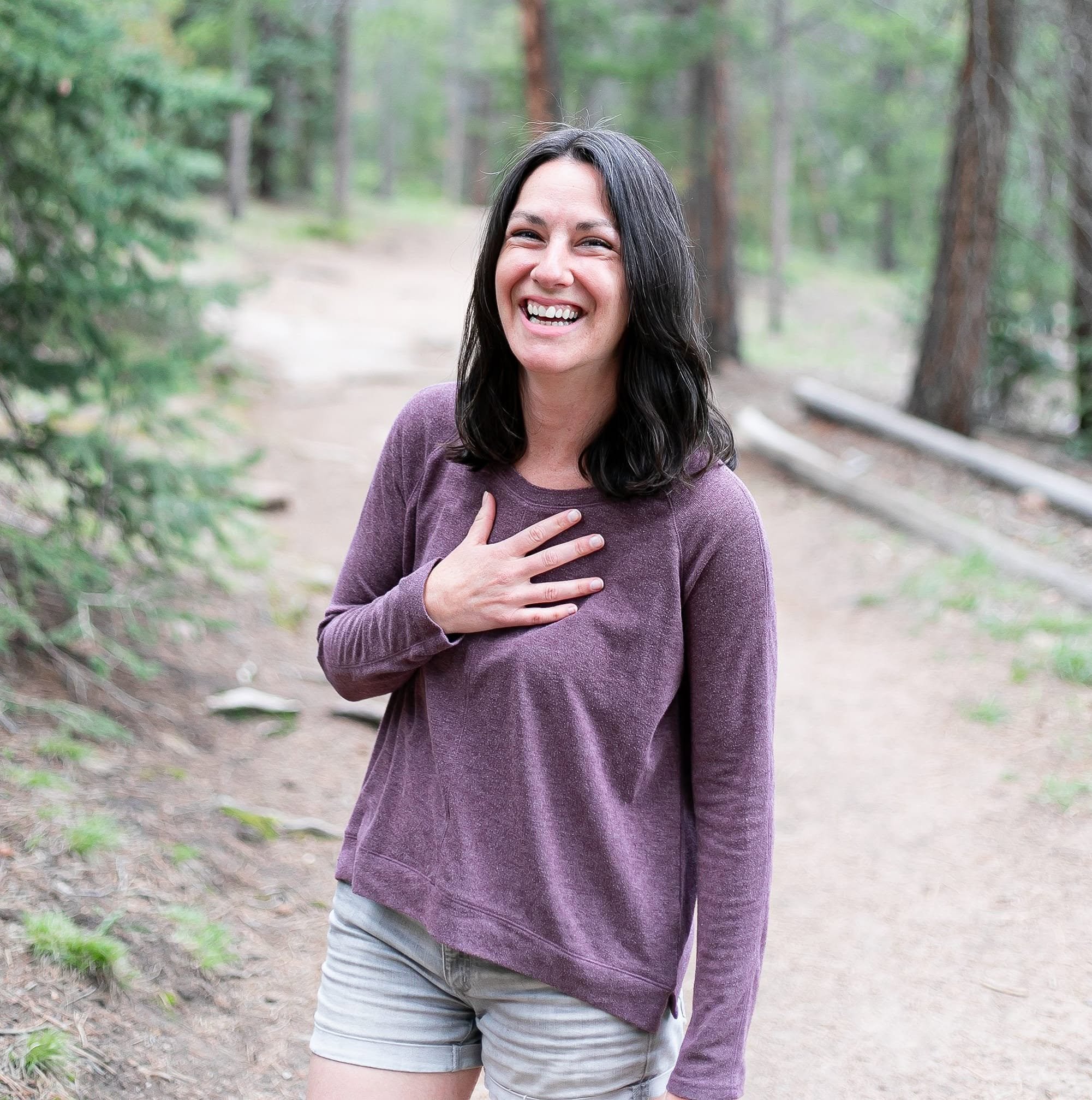 A woman with long dark hair, smiling, standing outdoors in a wooded area. She is wearing a purple long-sleeve shirt and khaki shorts, with her left hand on her chest. The background features a dirt path and green trees.