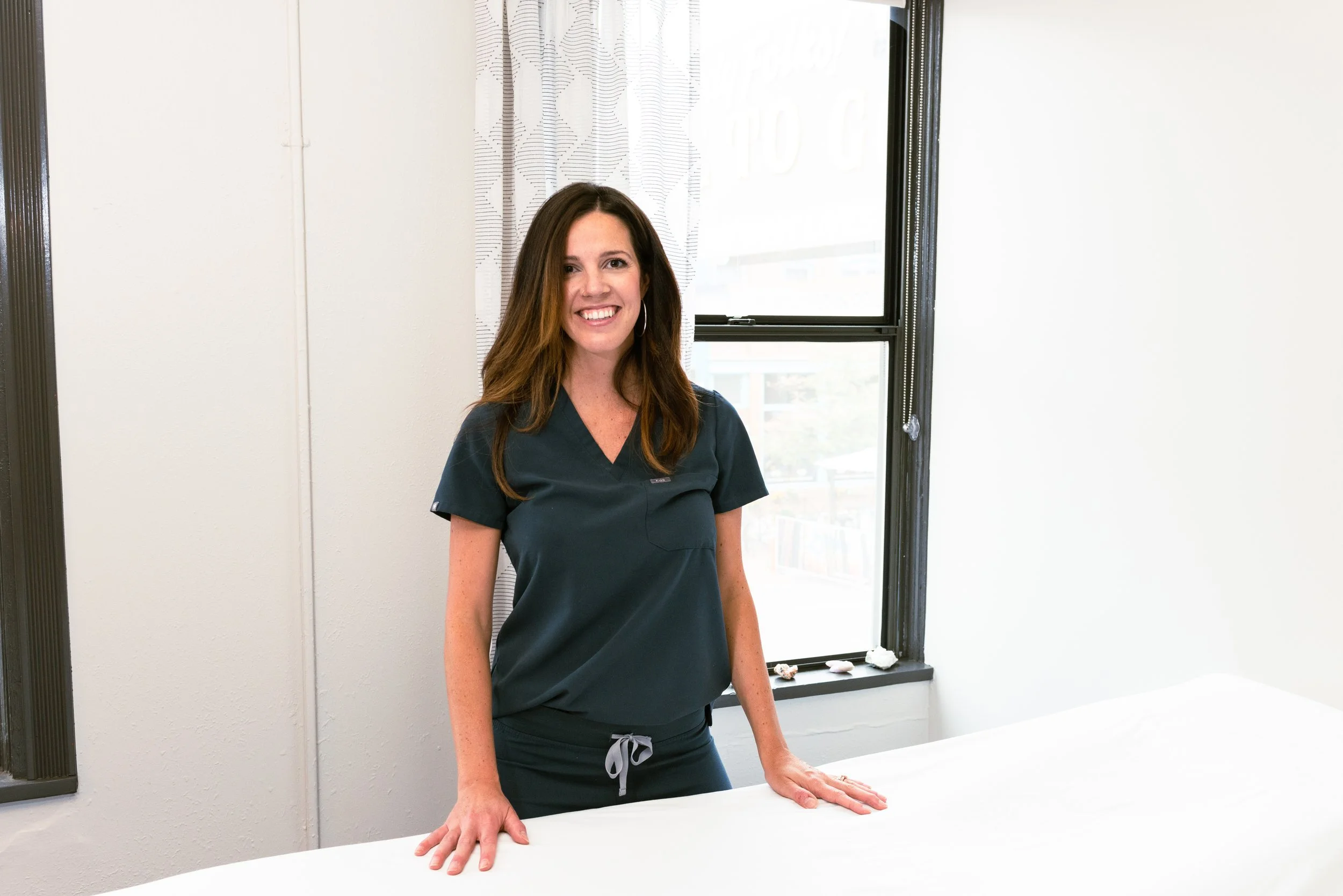 A woman with long brown hair, wearing scrubs, smiling and leaning on a white table inside a room with a window and curtain in the background.