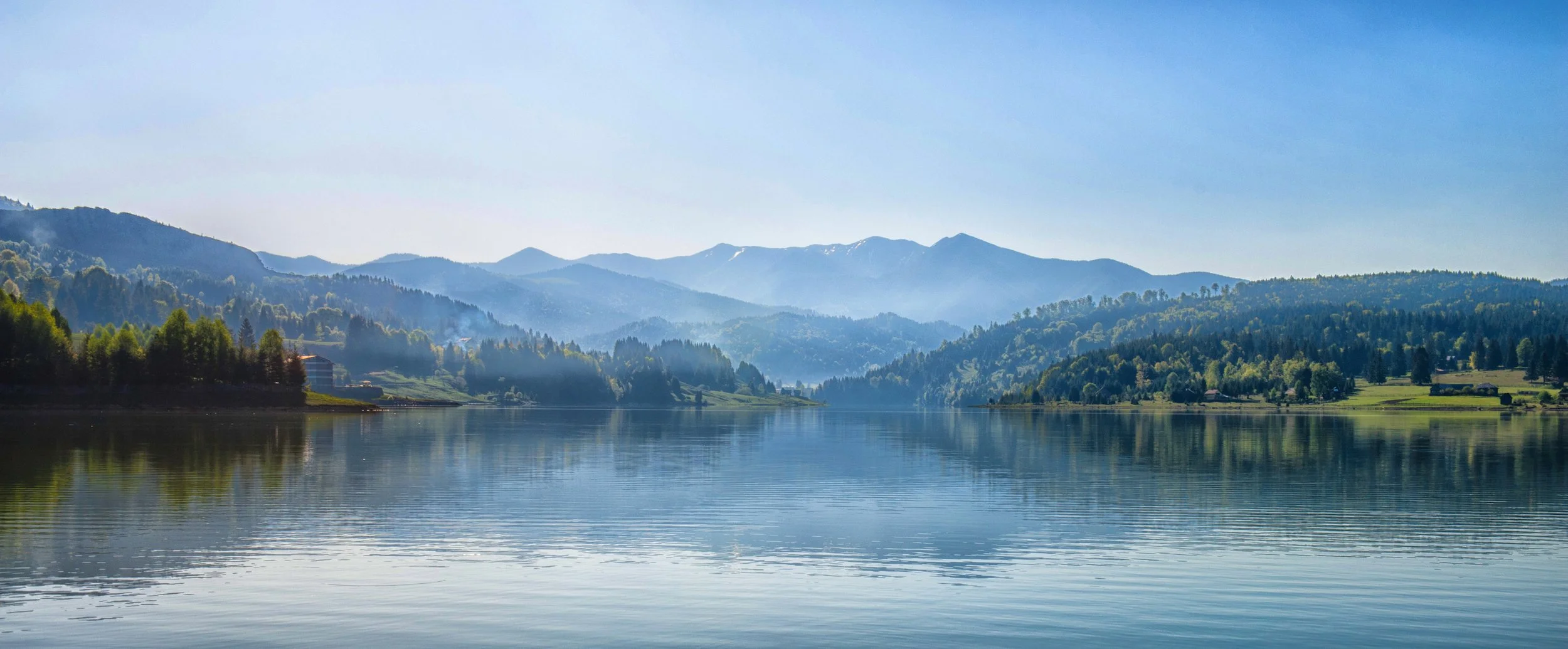 Calm lake with surrounding green forested hills and mountains in the distance under a clear blue sky.