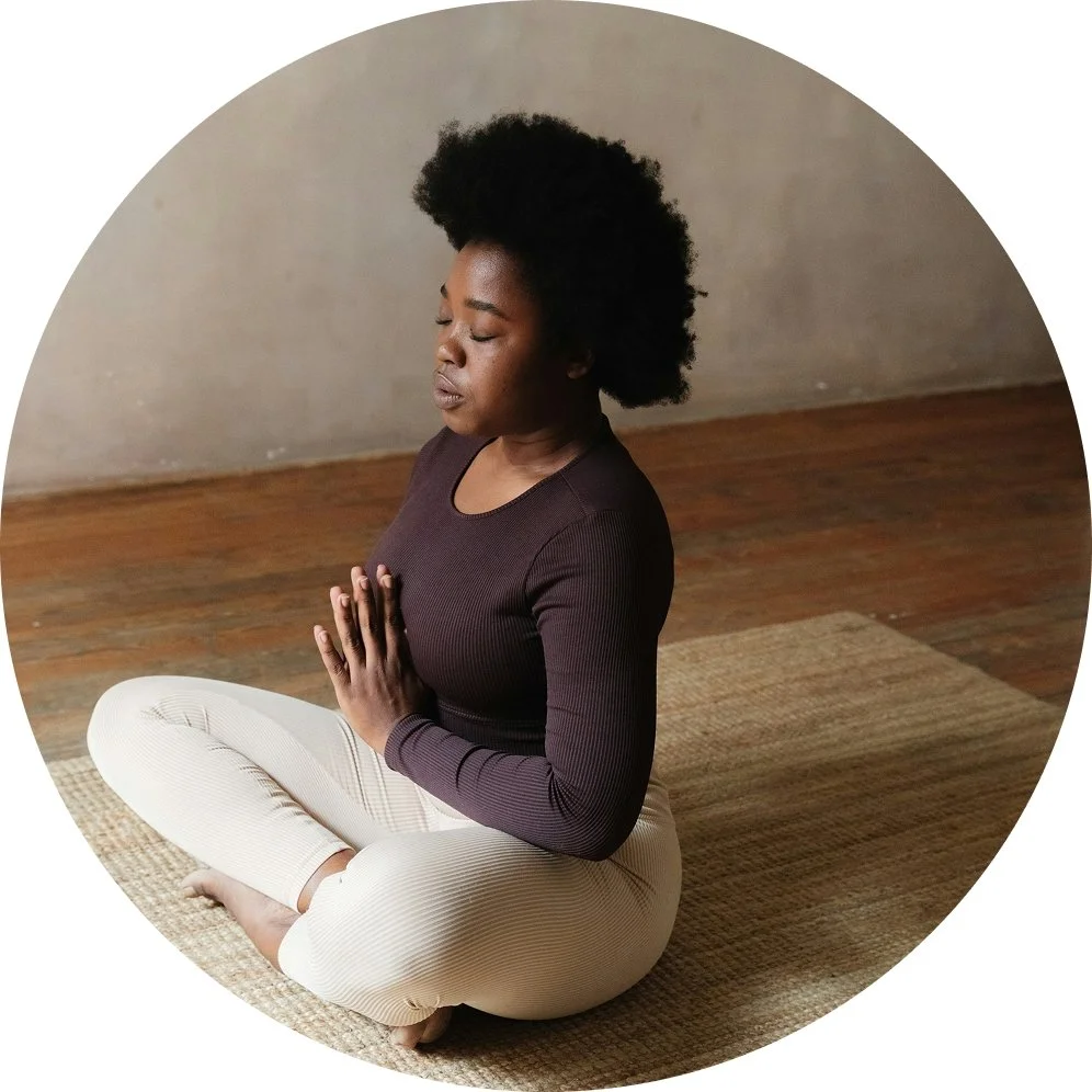 A woman practicing meditation or yoga indoors, sitting cross-legged on a mat with her hands in prayer position and eyes closed.