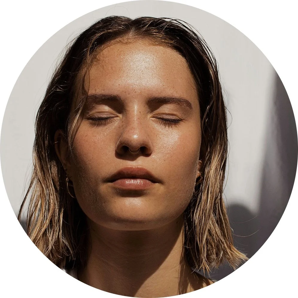 Close-up of a young woman with wet hair and closed eyes, lit by sunlight against a plain background.