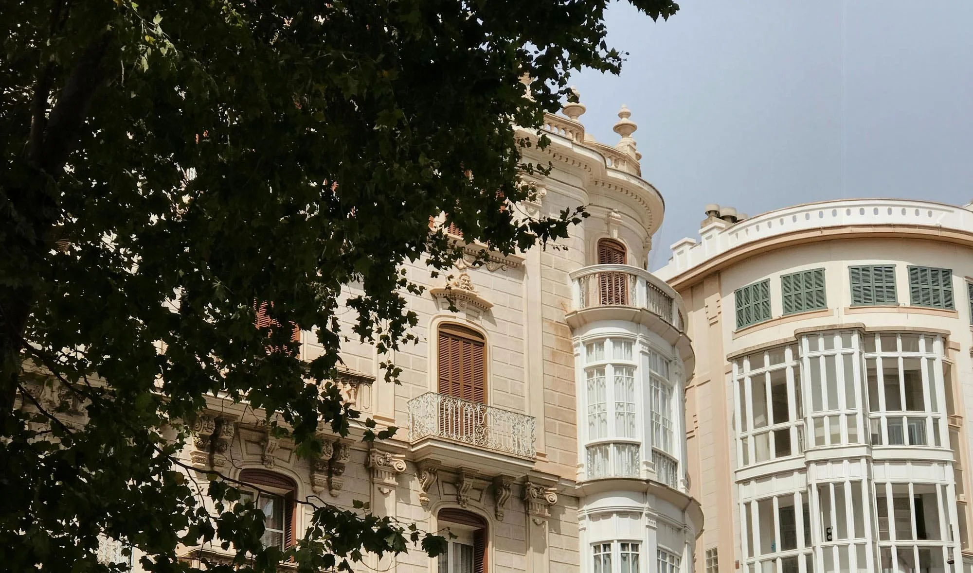 A view of ornate historic buildings with curved windows, balconies, and decorative architectural details, partially obscured by trees in the foreground.