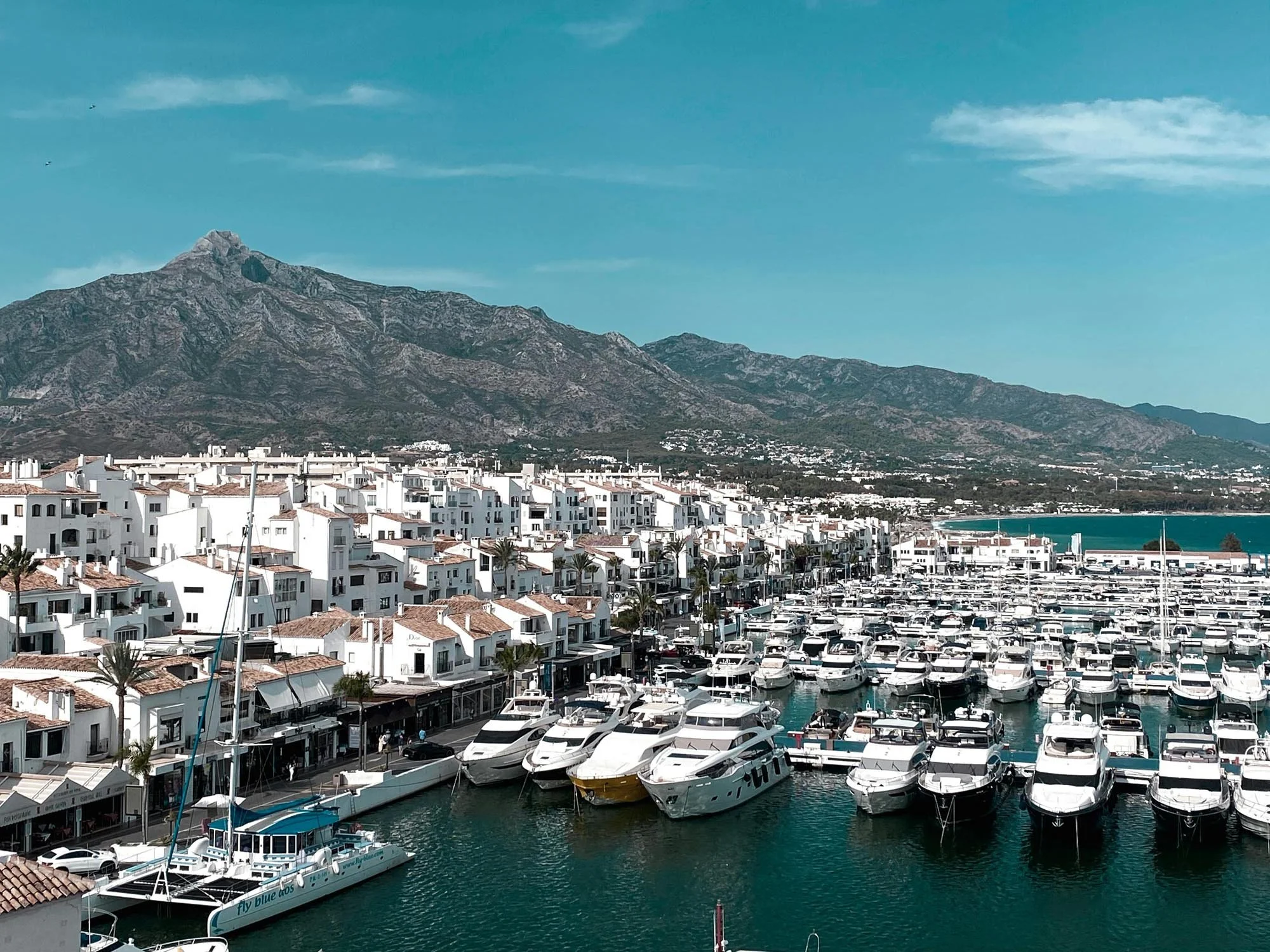 A marina filled with numerous white yachts and boats docked in the water, with white Mediterranean style buildings with terracotta roofs along the shoreline. In the background, a large mountain range extends across the horizon under a blue sky with some clouds.