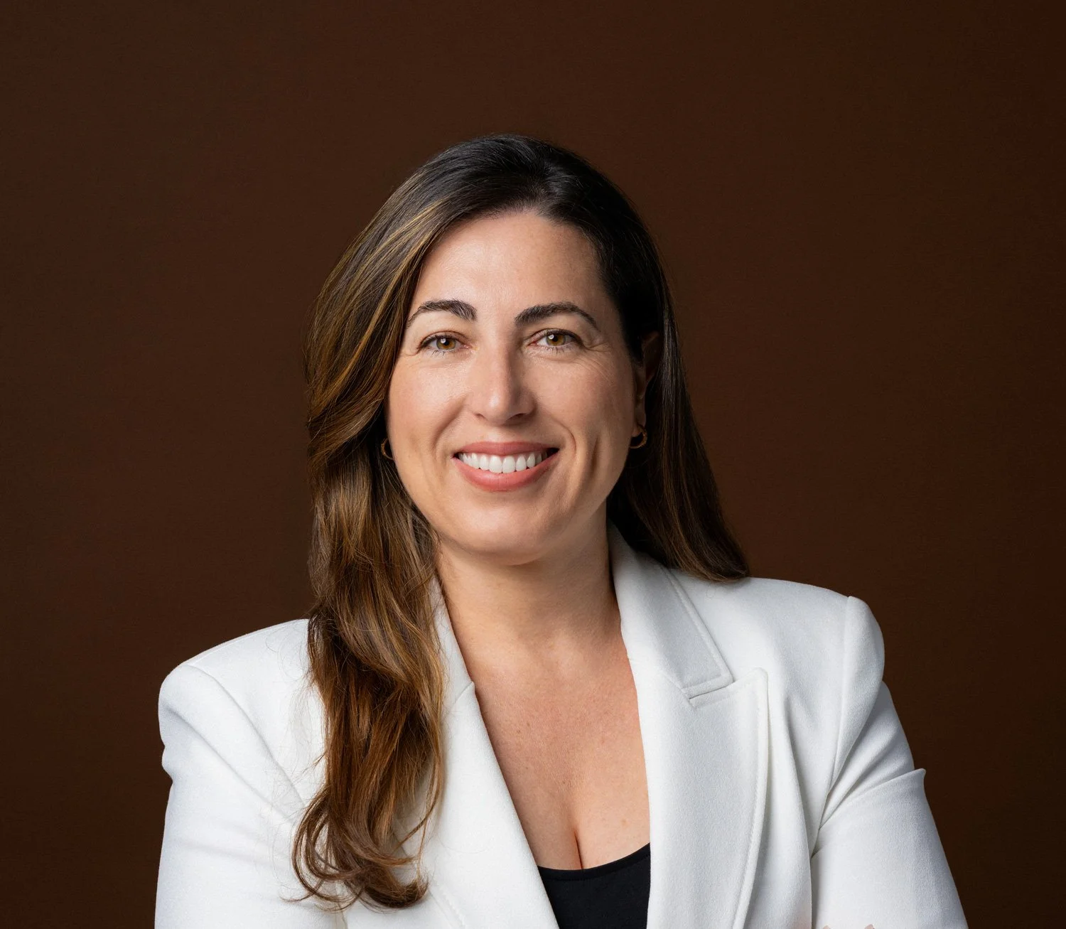 Ana Valencia, Founder of Altamare. Picture of a woman with long, wavy brown hair wearing a white blazer and a black top, smiling against a brown background.