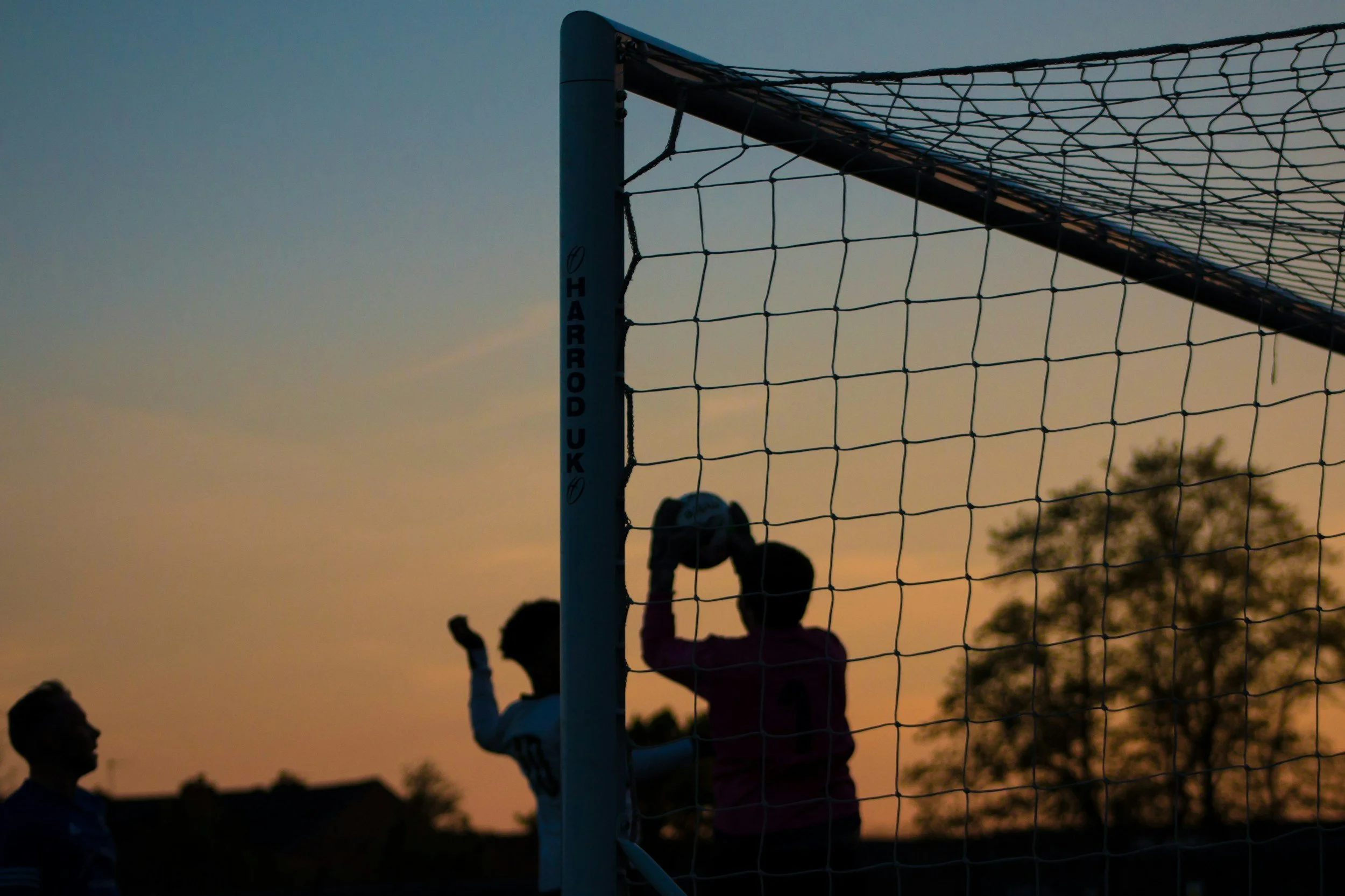 Fußballspiel bei Sonnenuntergang mit Kindern am Tor und Zuschauern im Hintergrund.