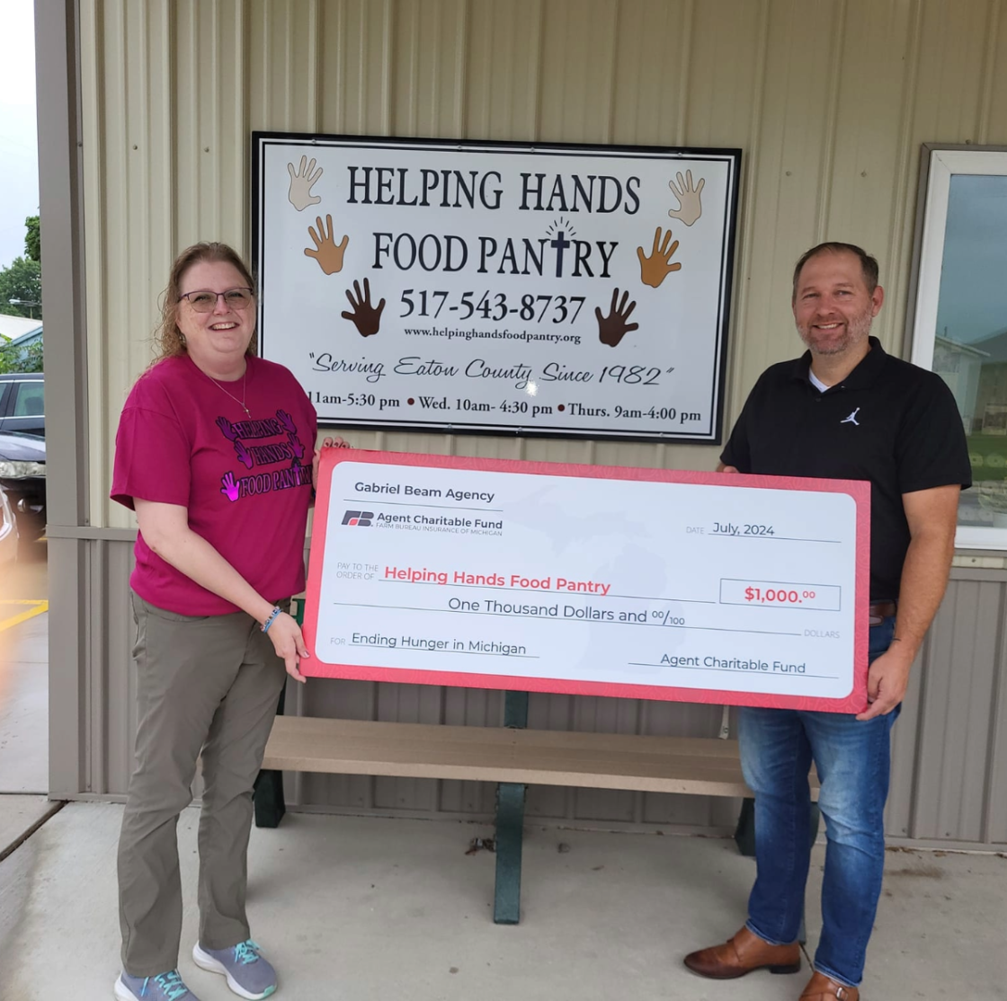 Two individuals holding a large check in front of a Helping Hands Food Pantry sign. The check is for $1,000 made out to Helping Hands Food Pantry, dated July 2024. The woman on the left wears glasses and a magenta shirt, the man on the right wears a black shirt and jeans.