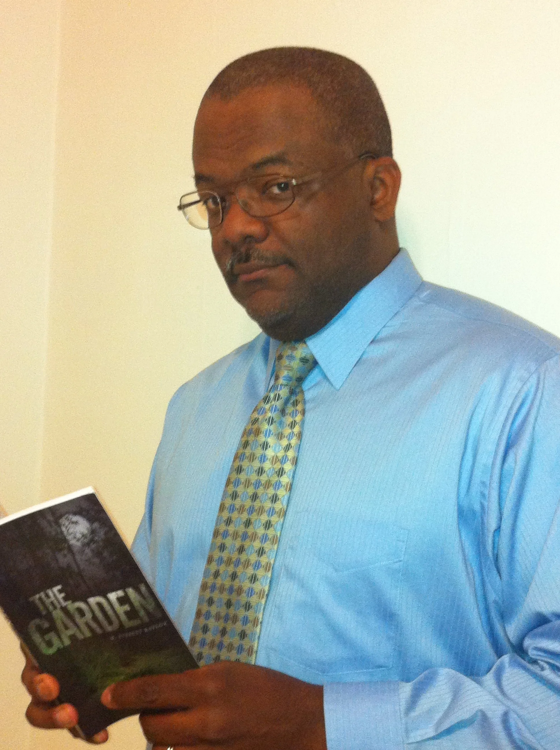 A man wearing glasses, a light blue shirt, and a patterned tie, holding a book titled 'The Garden' and standing against a plain wall.