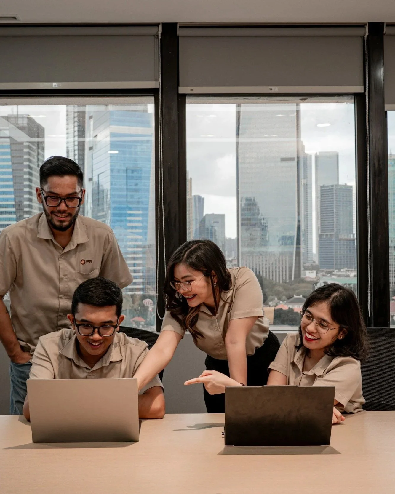 Five people in beige shirts looking at a laptop and discussing, with three women standing and two men sitting at a table, in a modern office with large windows showing a city skyline.