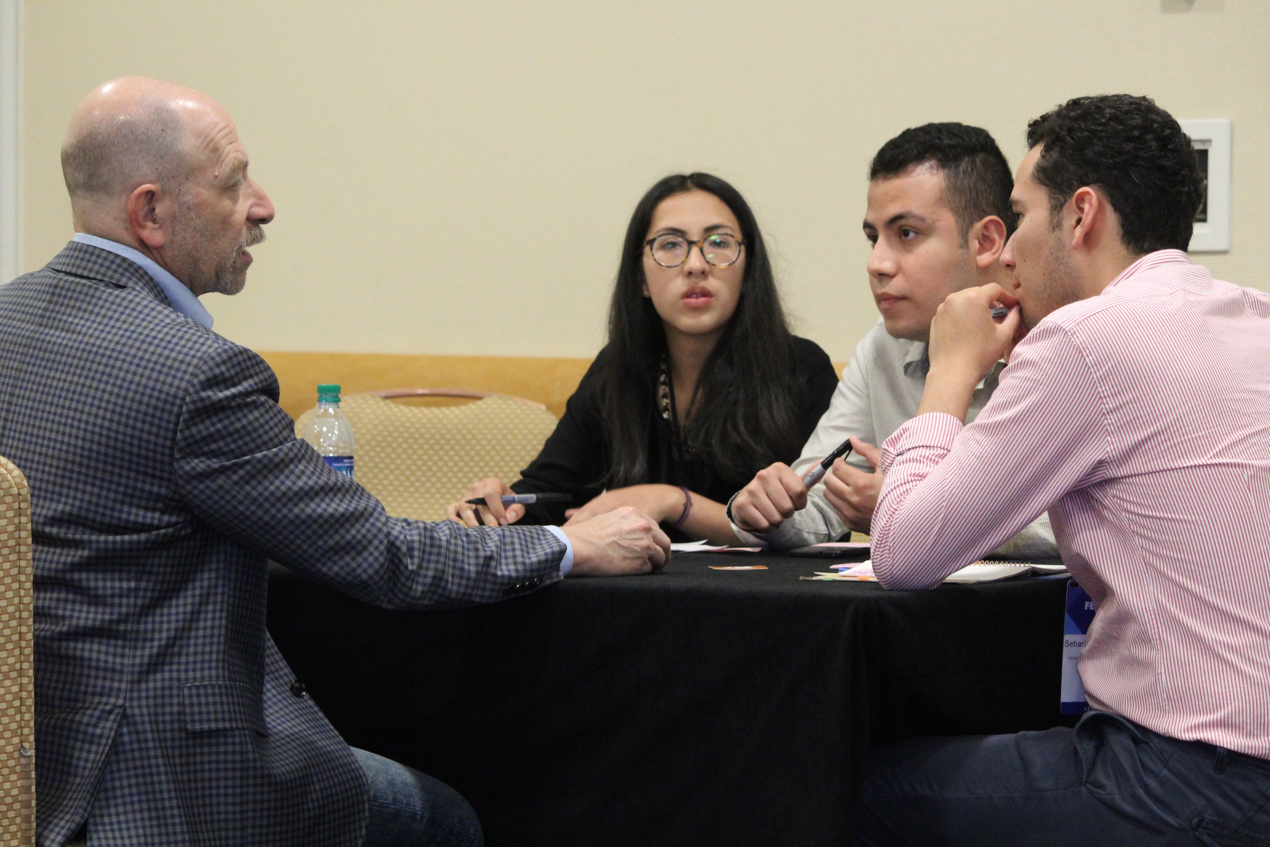 A group of four diverse people is engaged in a serious discussion at a conference or workshop, sitting around a table with notes and pens.
