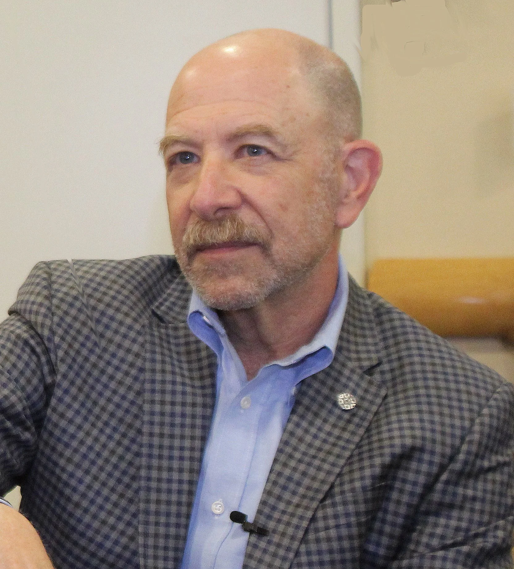 Close-up of a middle-aged man with a shaved head, gray beard, and blue eyes, dressed in a checkered blazer, light blue shirt, and a pin on his blazer, sitting indoors.