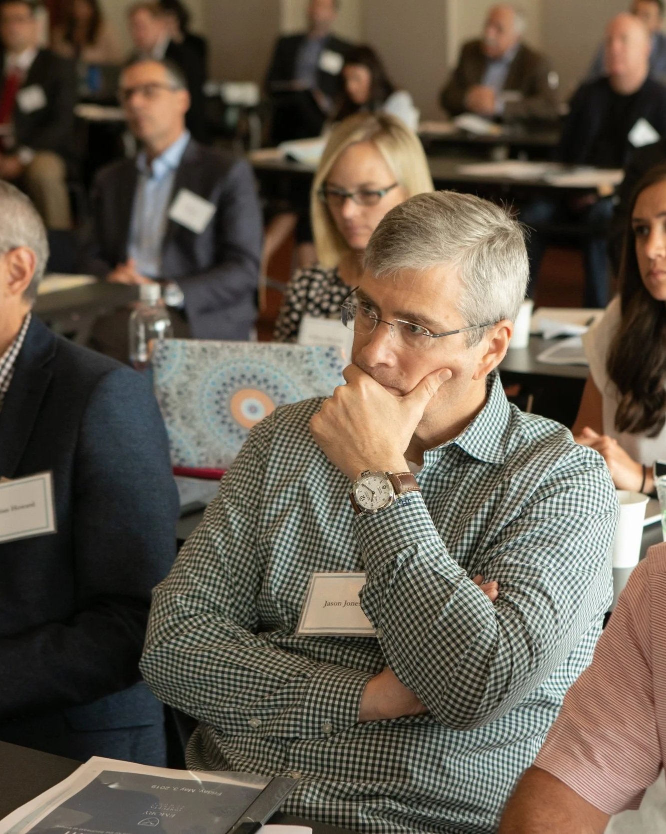 A group of professionally dressed people attending a conference or seminar in a conference room, seated at tables, with some taking notes and listening attentively.