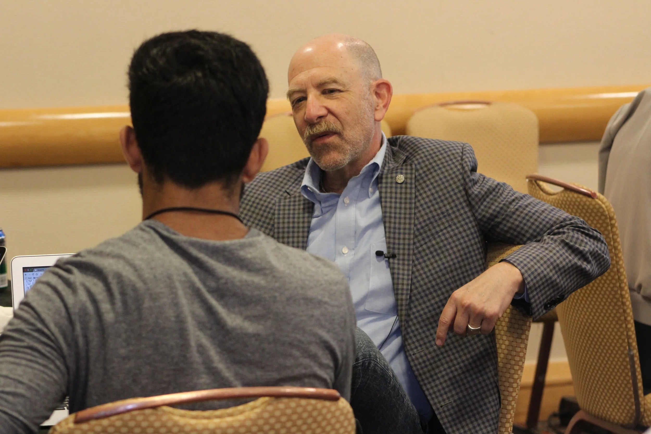 Two men seated and engaged in conversation in a conference room, with one man facing away from the camera and the other facing towards the camera, dressed in a checked blazer.