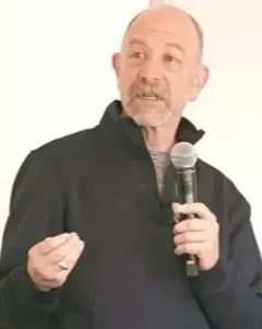 A man speaking into a microphone, wearing a black jacket, with a white background.