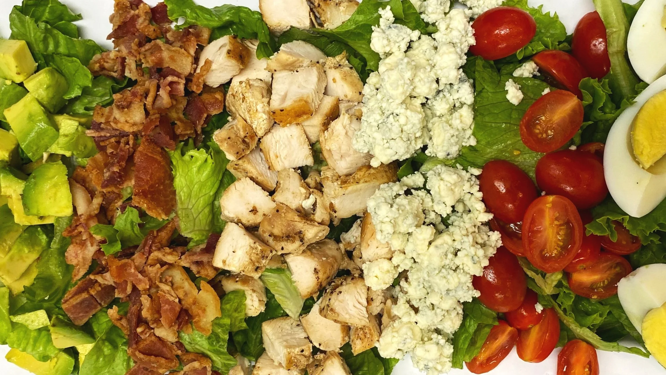 A mixed salad with chopped lettuce, diced avocado, bacon bits, grilled chicken pieces, crumbled blue cheese, cherry tomatoes, and sliced hard-boiled eggs.