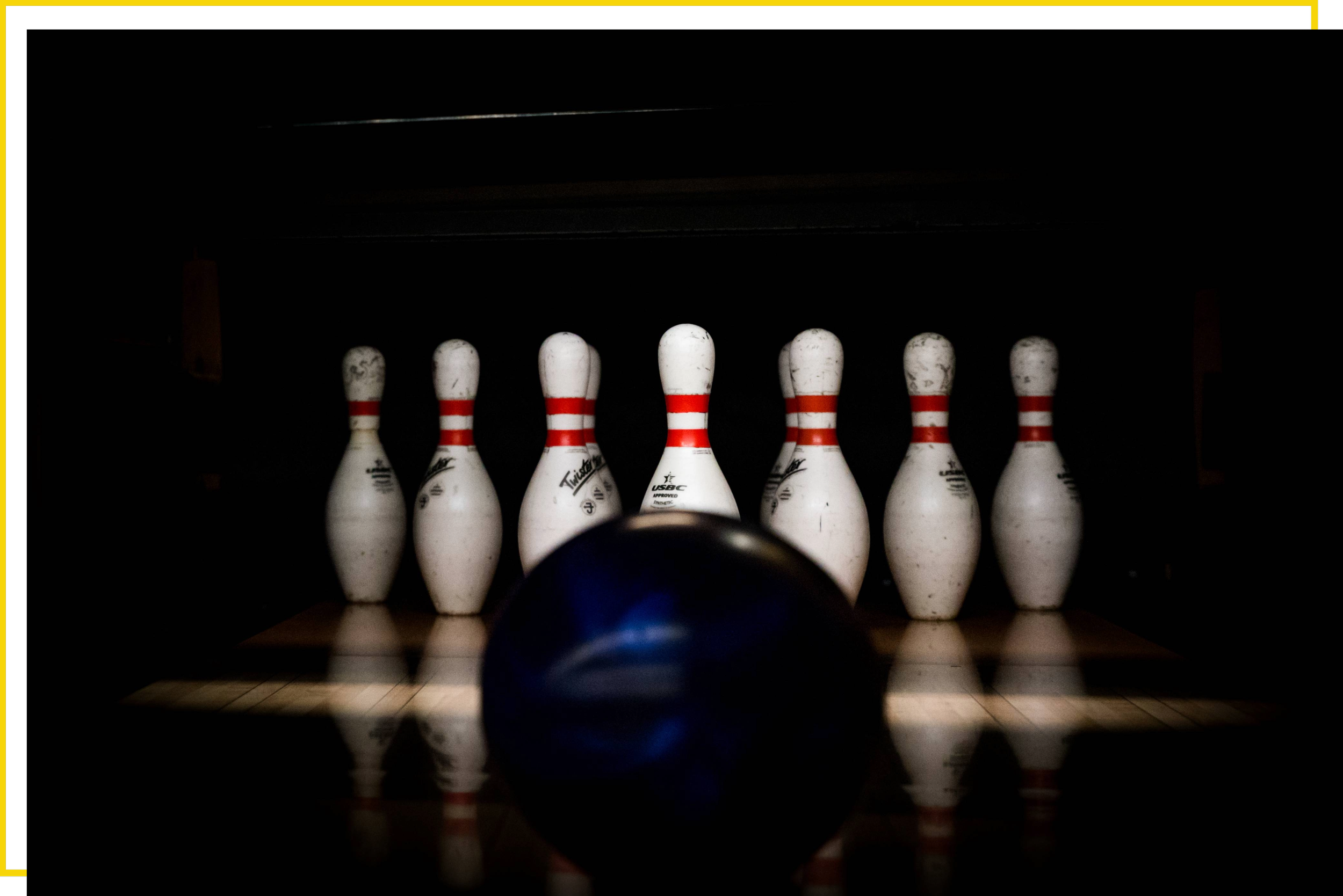 A black bowling ball in the foreground facing seven white bowling pins with red stripes, set up on a bowling alley with dark surroundings.
