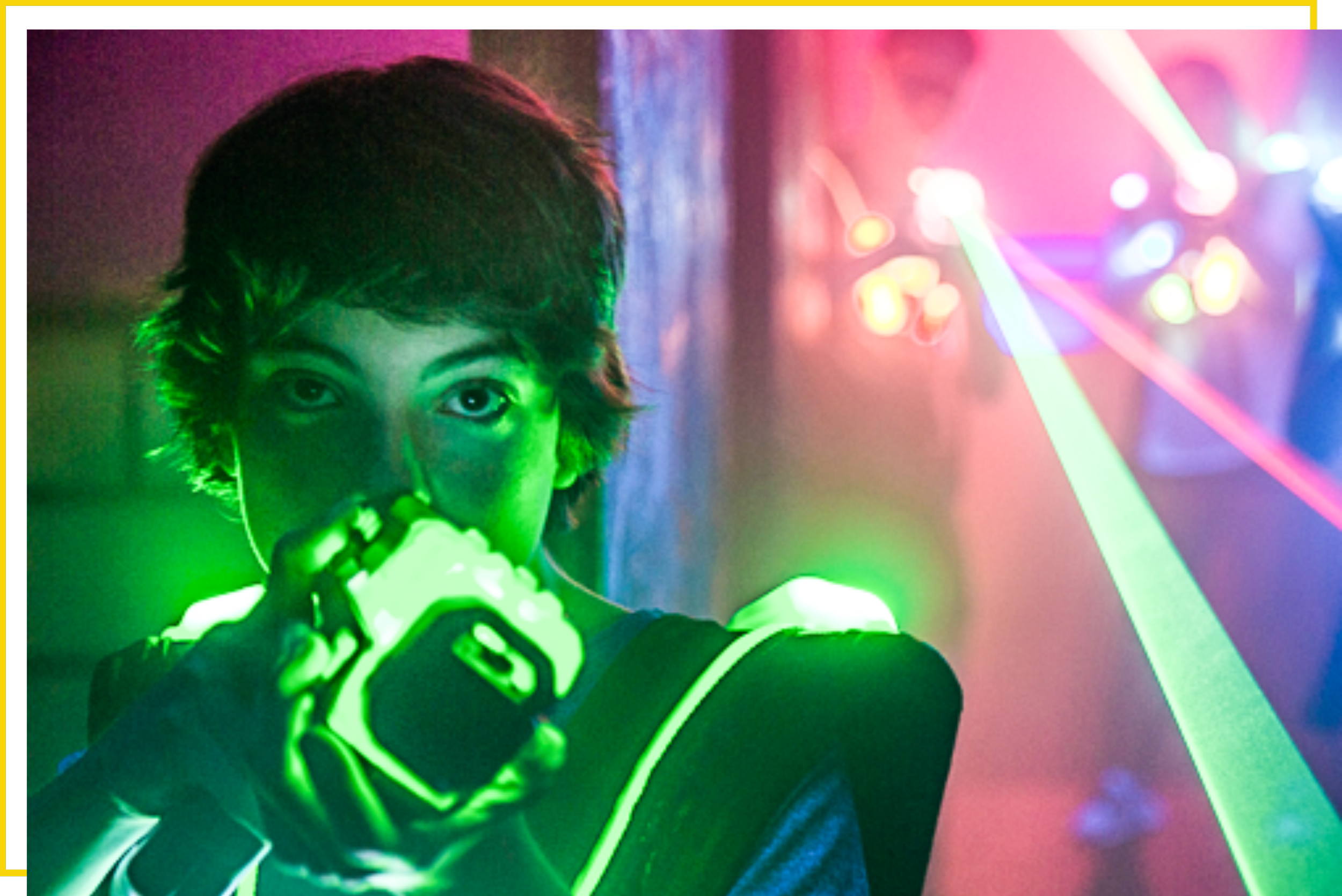 A young person with short hair holding a glow-in-the-dark laser tag device, illuminated by green light, in a room with colorful laser lights projecting on the wall while playing laser tag.