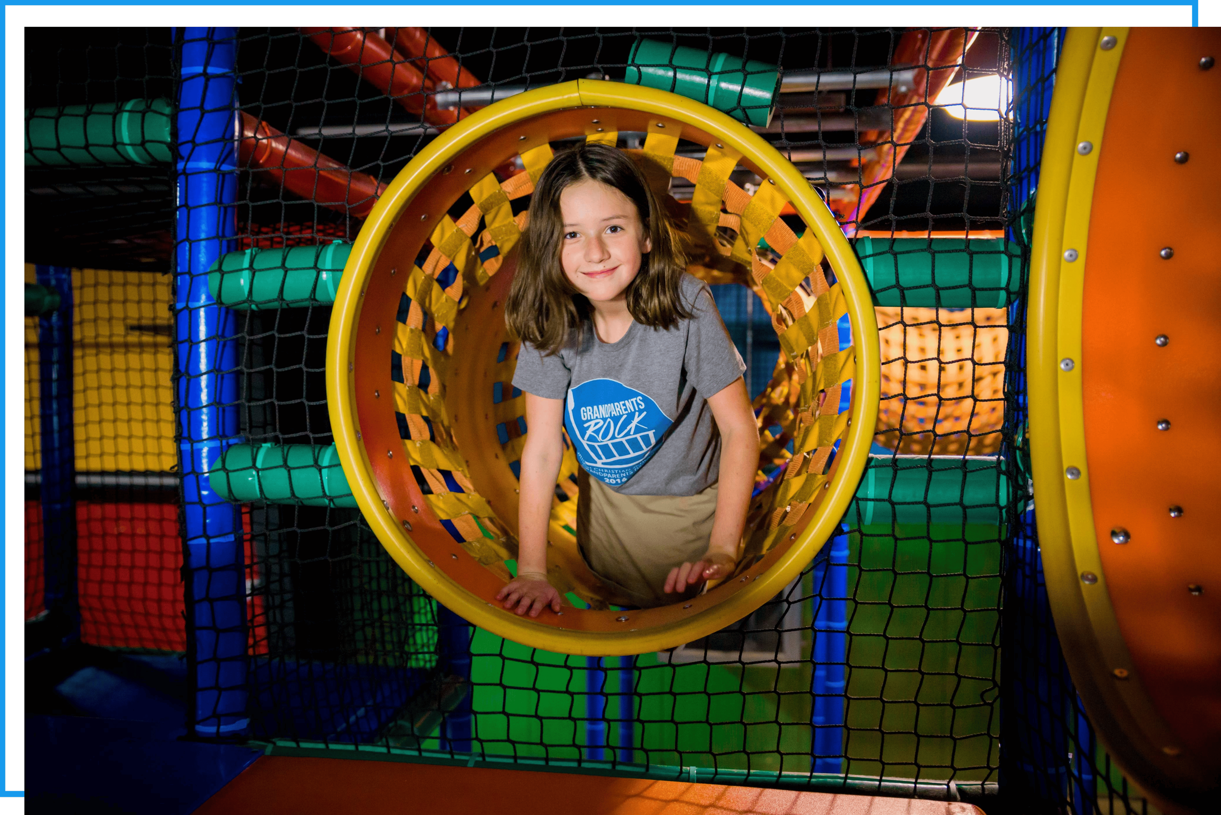 A young girl with brown hair smiling in a tunnel at an indoor play area, surrounded by colorful play structures and netting.