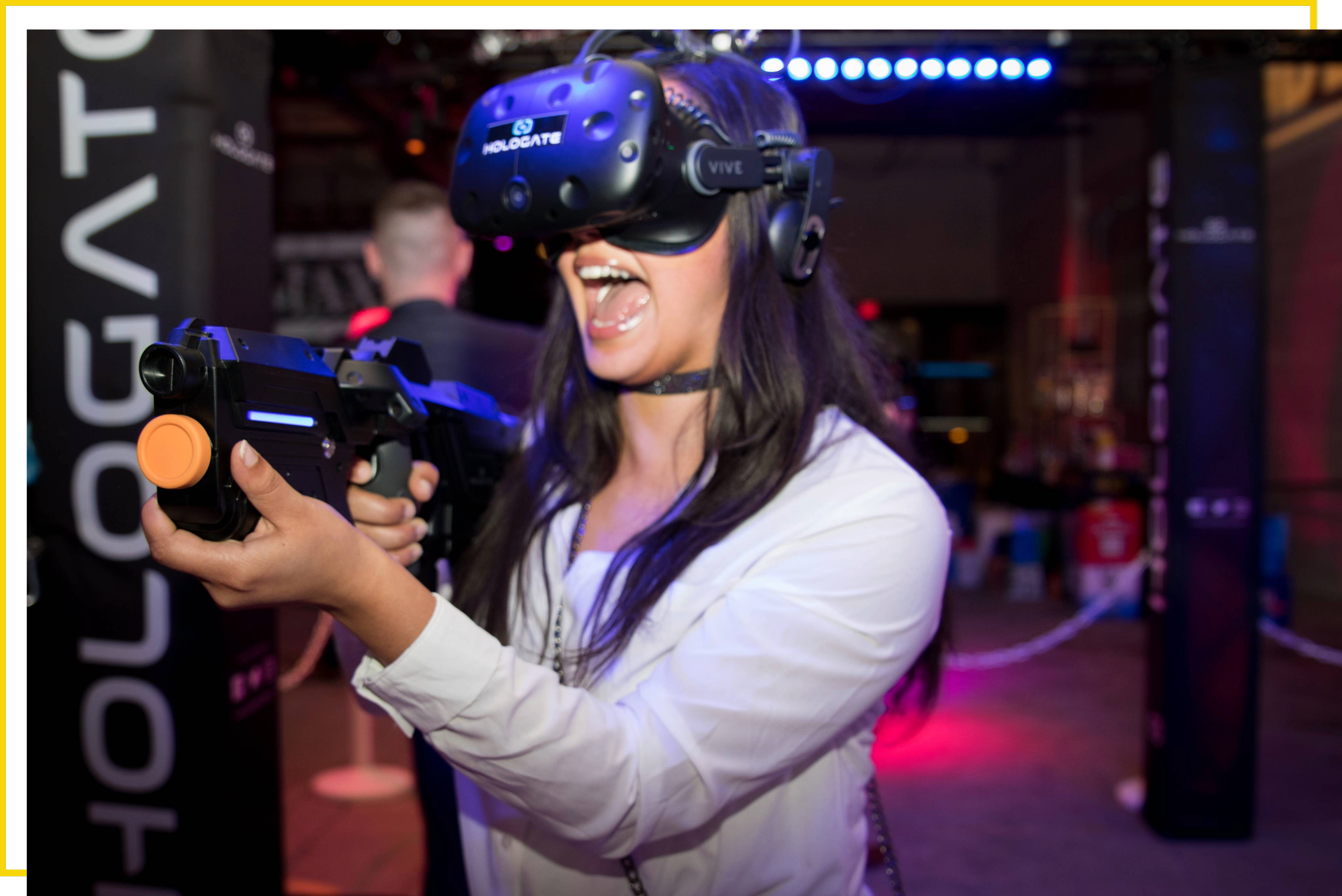 A woman wearing a virtual reality headset and holding a VR gun controller, excited and smiling at a gaming event.