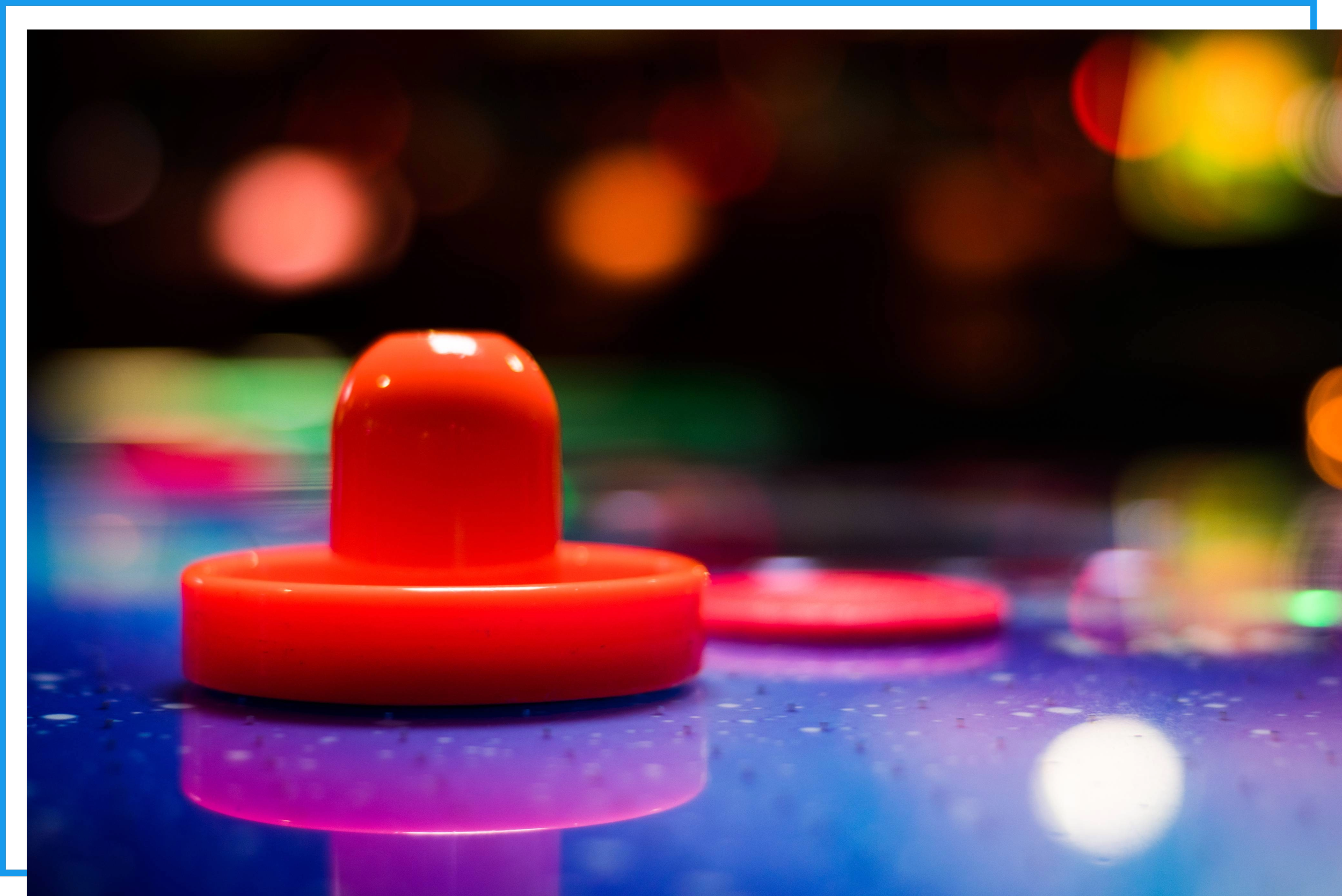 Close-up of an air hockey table. It's sitting on a reflective surface with a blurred, colorful background of bokeh lights.