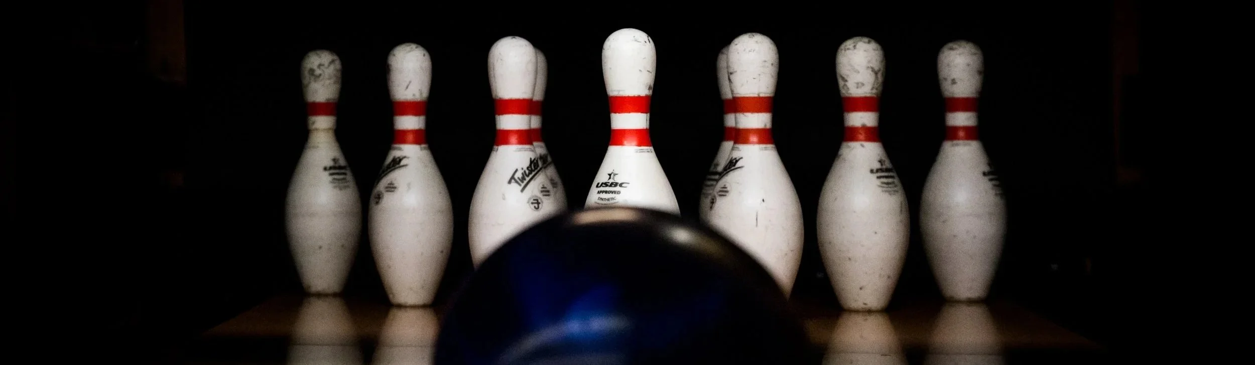 Seven white bowling pins with red stripes, arranged in a row behind a black bowling ball on a lane.