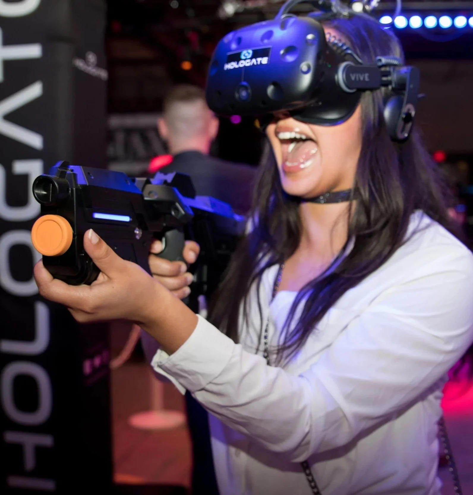 A woman with dark hair wearing a white shirt and a choker, excitedly playing a virtual reality shooting game with a VR headset and a gun controller in a lively, colorful environment.